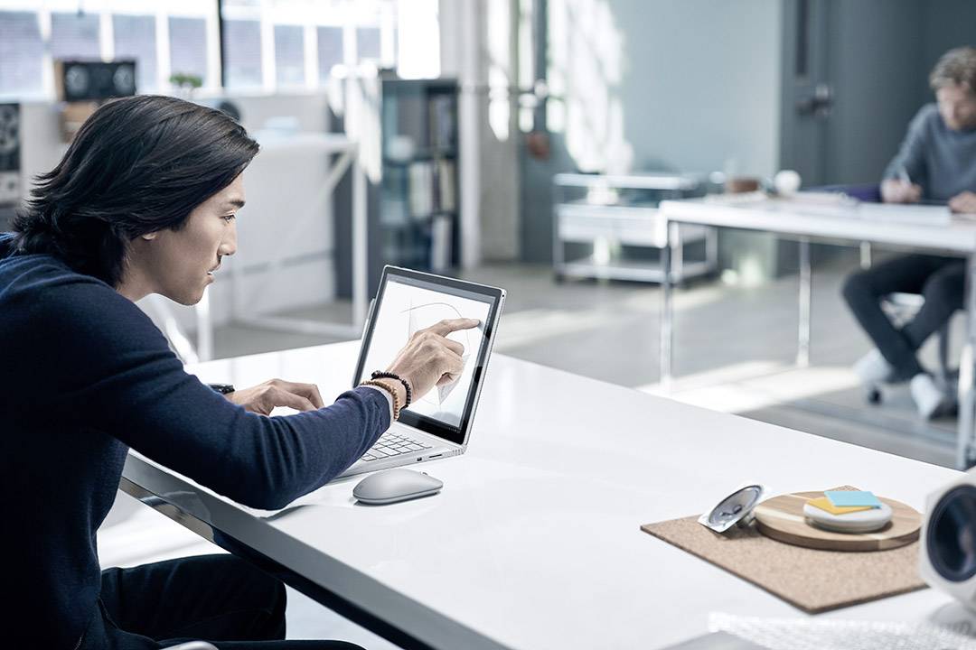 Un homme est assis à un bureau et travaille avec un stylet numérique sur un ordinateur portable. Un bureau est visible en arrière-plan.