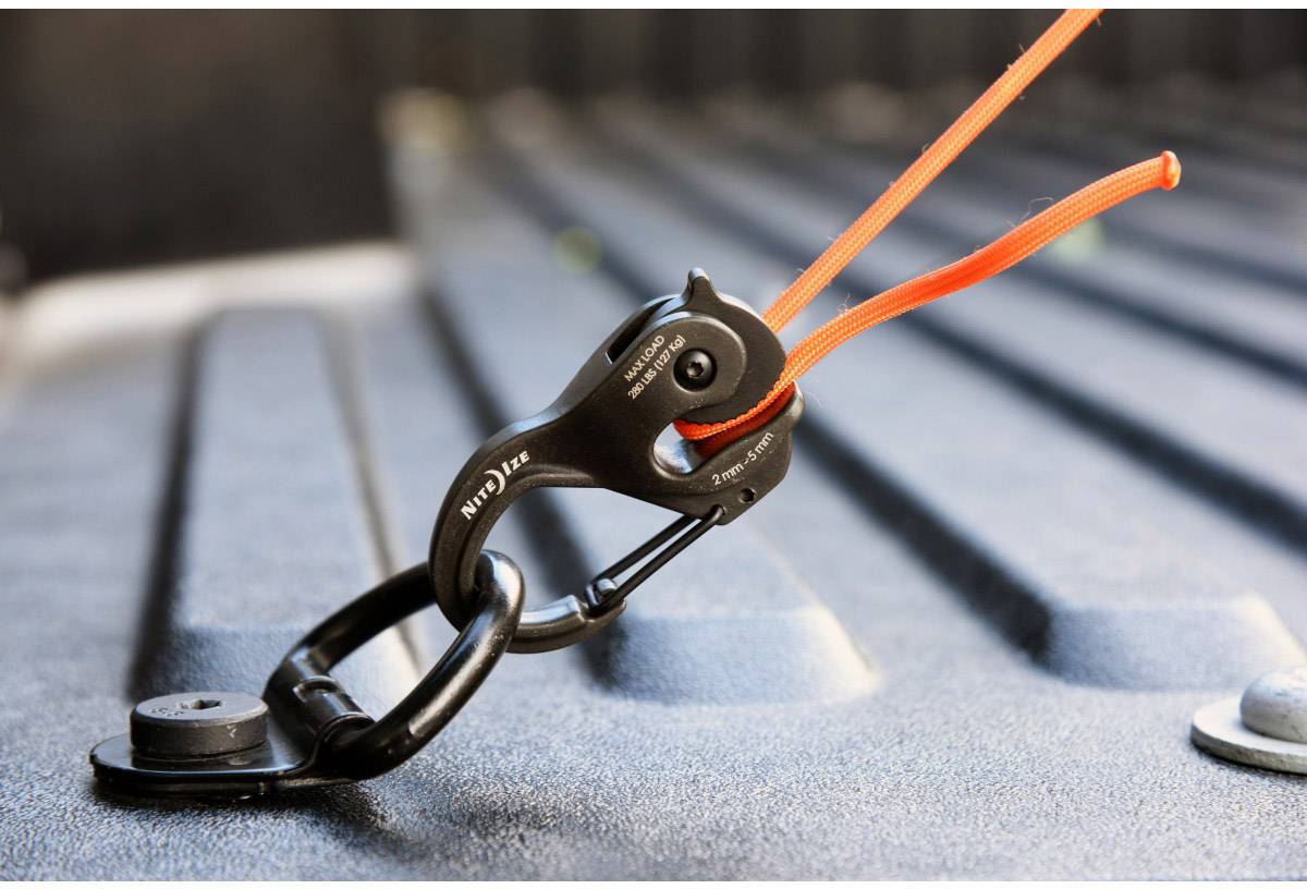Un mousqueton noir sécurise une corde orange sur une surface métallique, probablement pour l'arrimage de la charge.