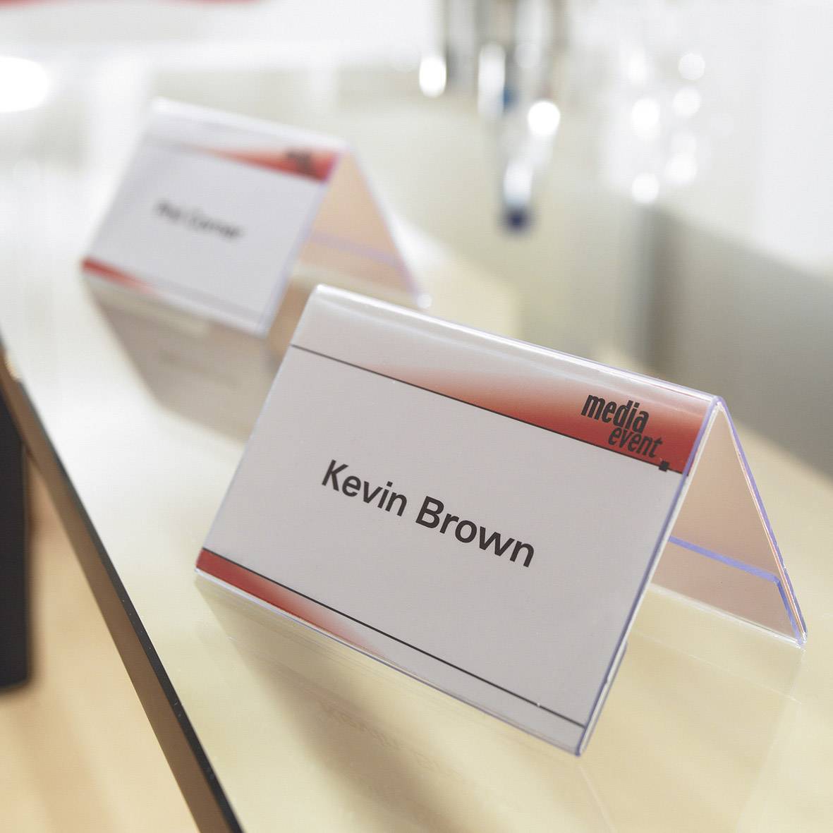 Plaque nominative portant l'inscription 'Kevin Brown' sur une table lors d'un événement médiatique, avec l'accent mis sur la plaque et un arrière-plan flou.