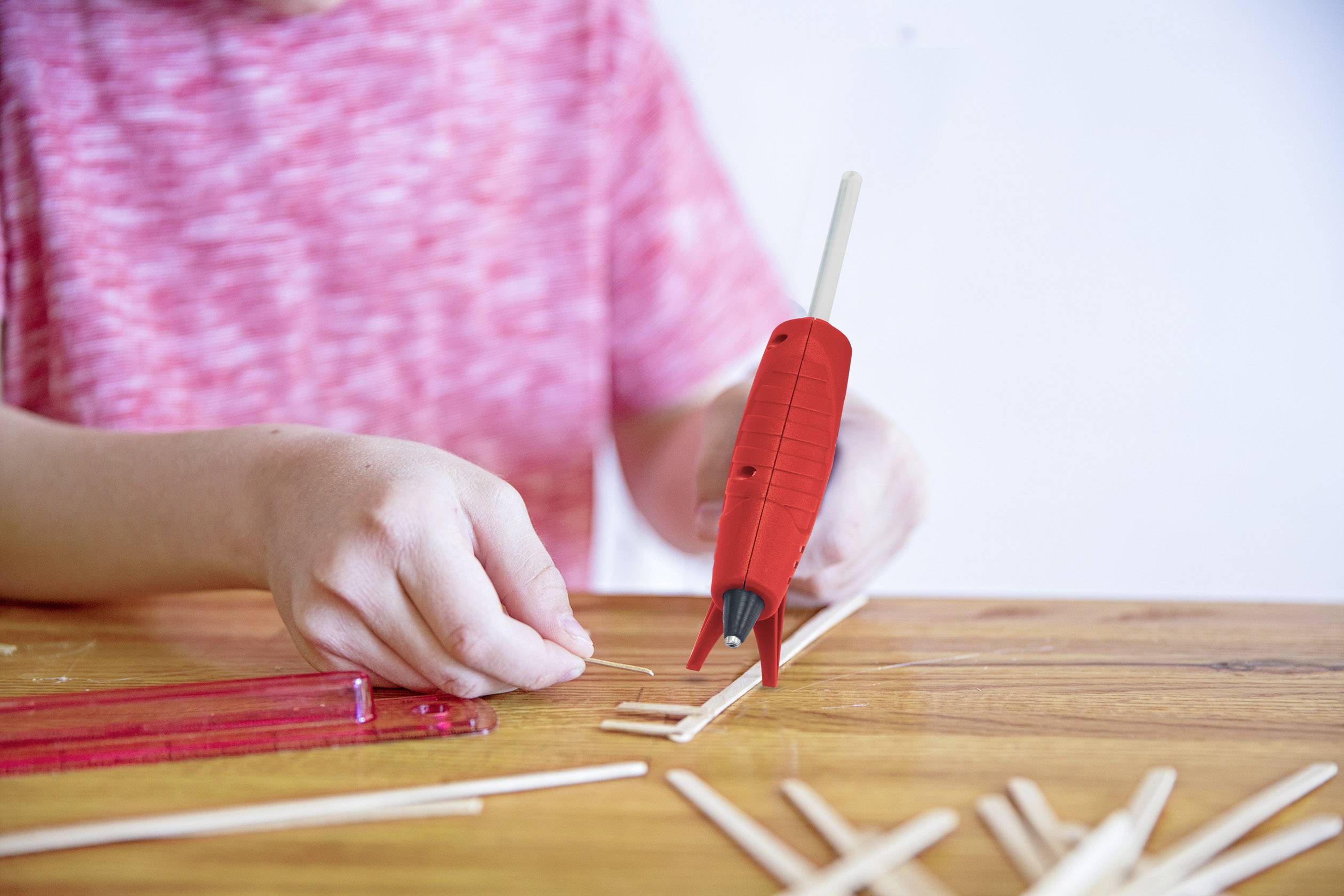 Un enfant construit avec des bâtonnets en bois et un pistolet à colle chaude sur une table en bois. Il porte un t-shirt rouge rayé.
