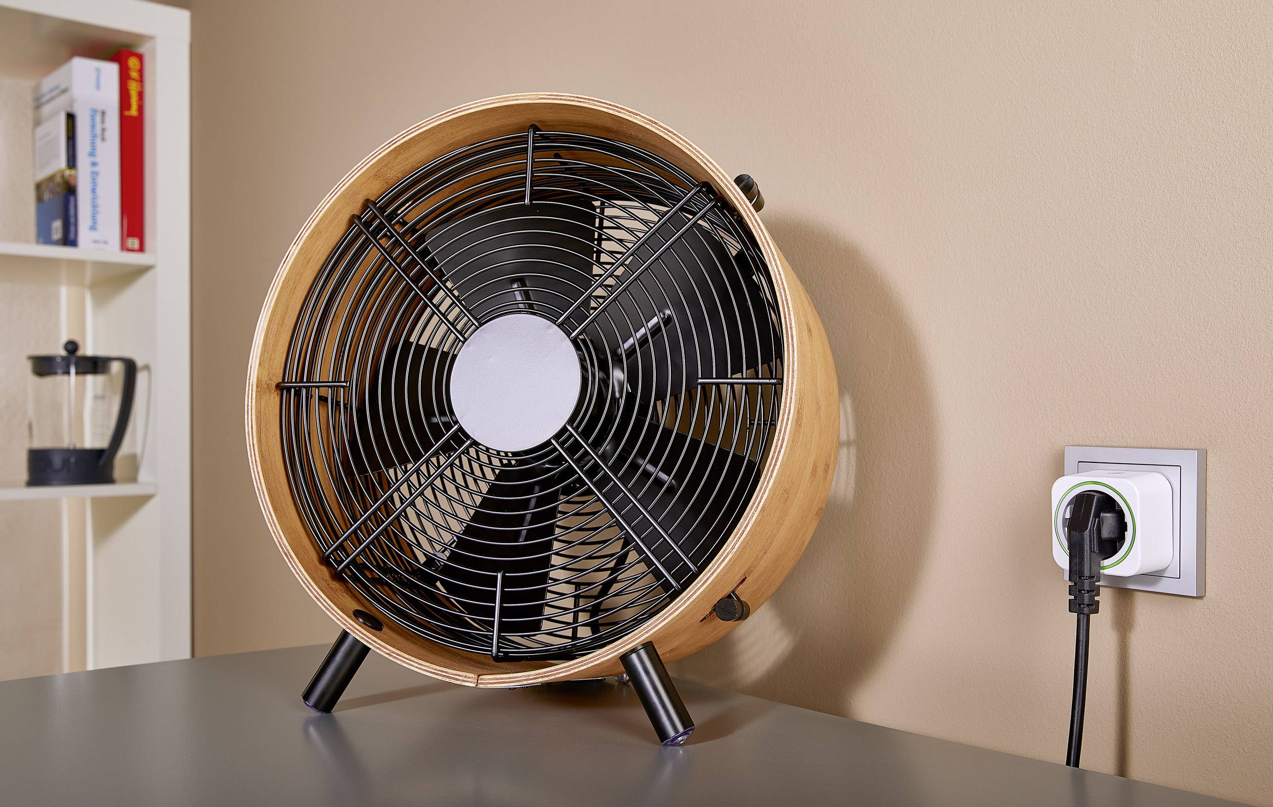 Un ventilateur de table au design rétro est posé sur une table grise. Il est branché et prêt à être utilisé. En arrière-plan, une bibliothèque.