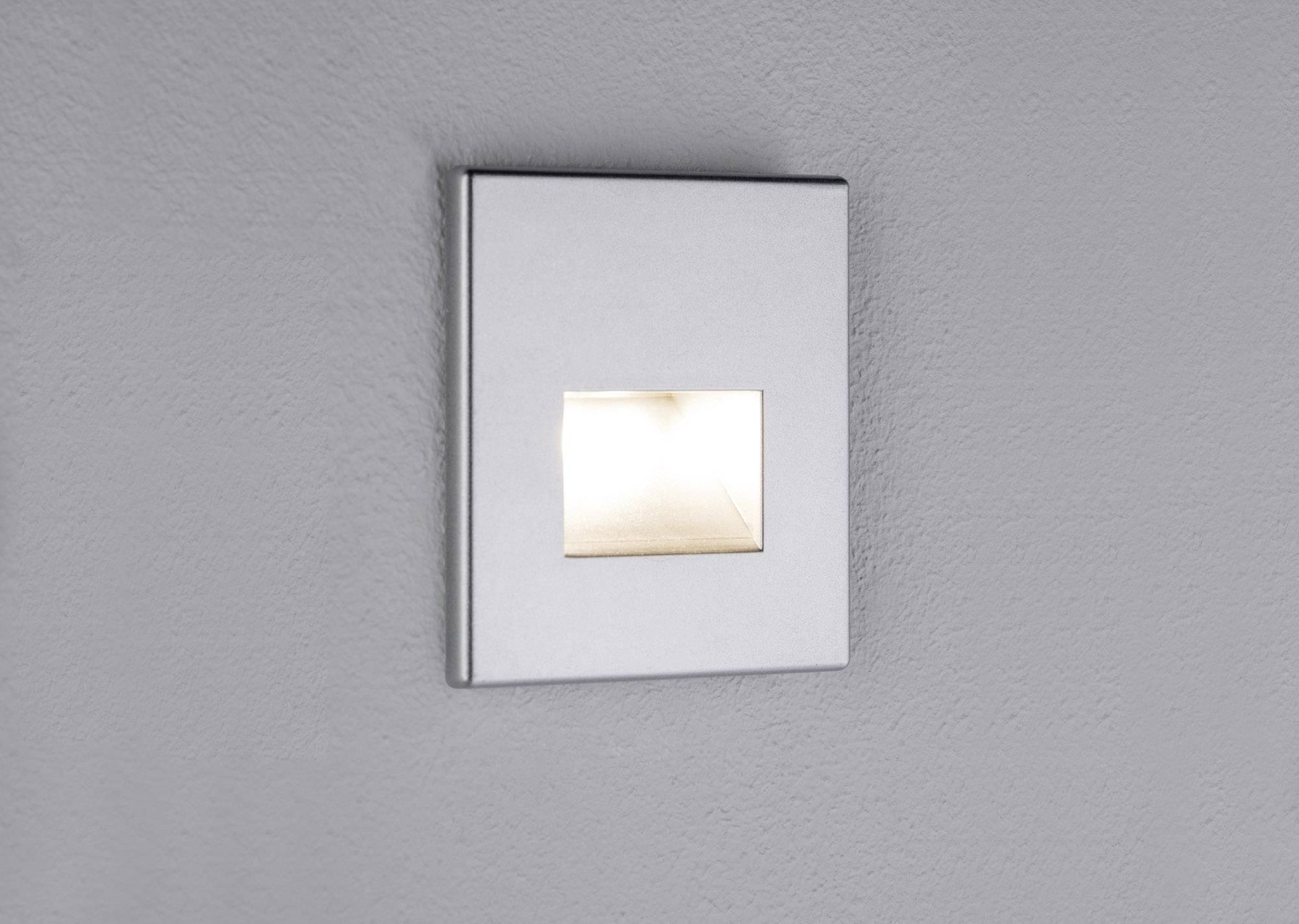 Un applique murale rectangulaire argentée diffuse une lumière douce sur un mur gris, sans autres éléments décoratifs.