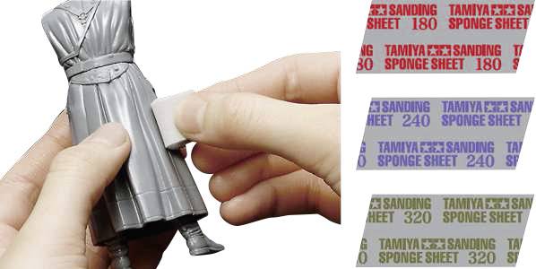 La main ponce une figurine avec une éponge de ponçage ; à droite, papiers de verre de différentes grains 180, 240, 320 de Tamiya