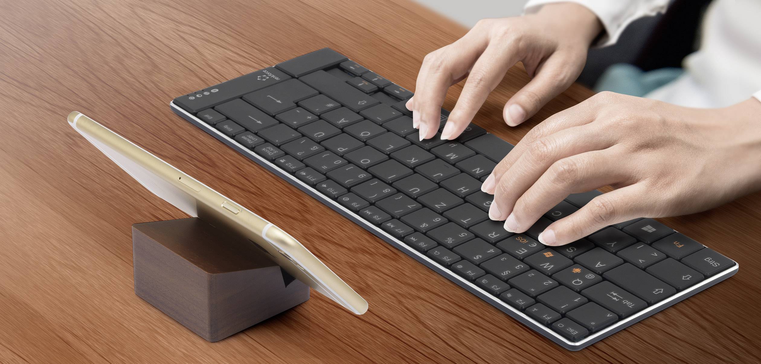 Une personne tape sur un clavier noir sans fil. Un smartphone doré est posé à côté d'elle sur un support en bois.