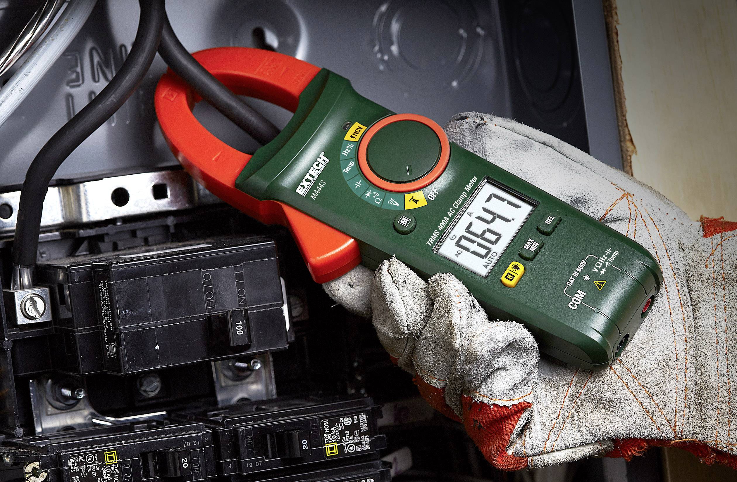 Une personne tient un multimètre vert contre un composant électrique et mesure une tension de 164,7 volts. L'appareil affiche différents modes de mesure.