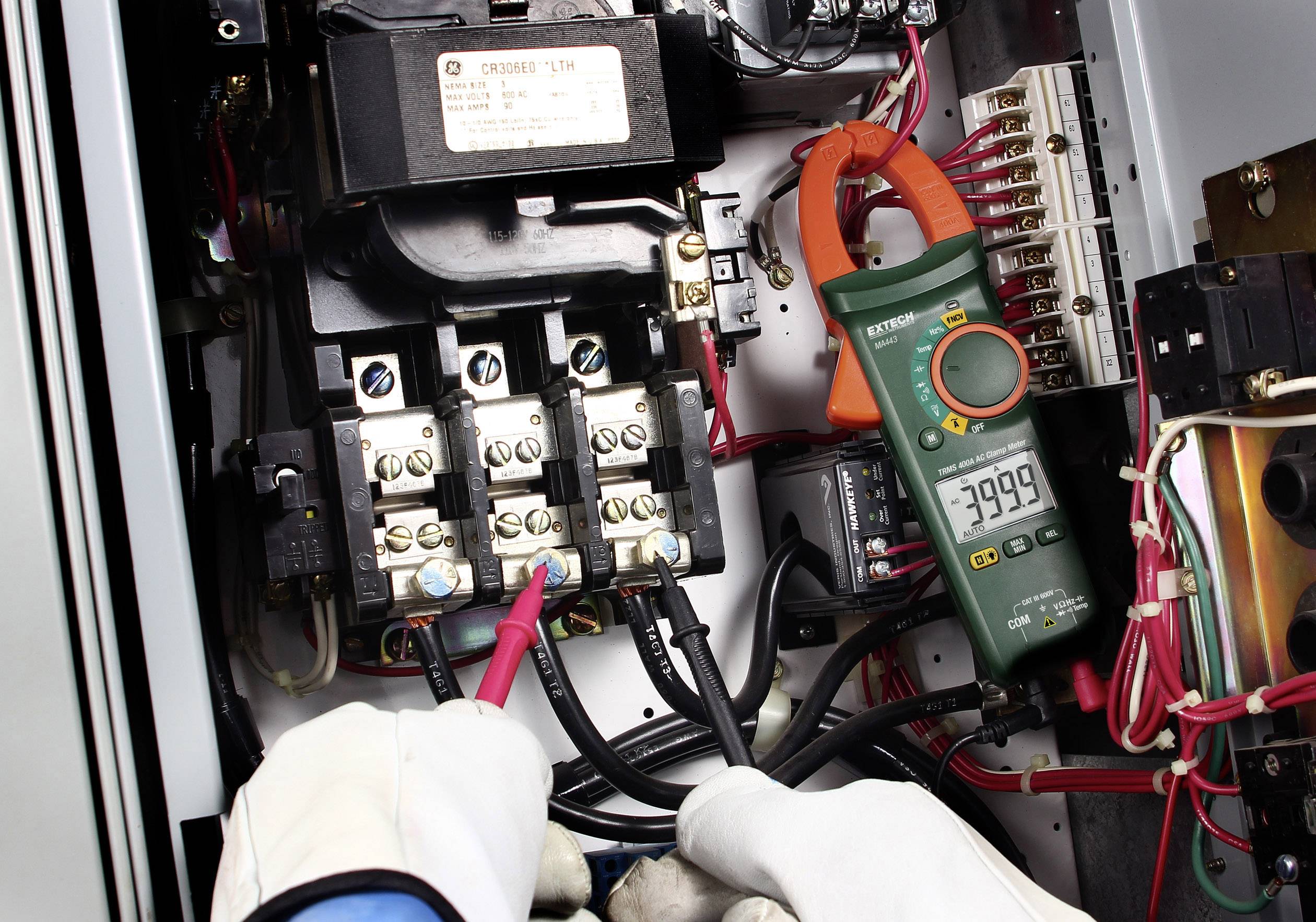 Un électricien mesure la tension électrique dans un tableau électrique à l'aide d'un multimètre, affichant une valeur de -3999. Des gants le protègent contre les chocs électriques.