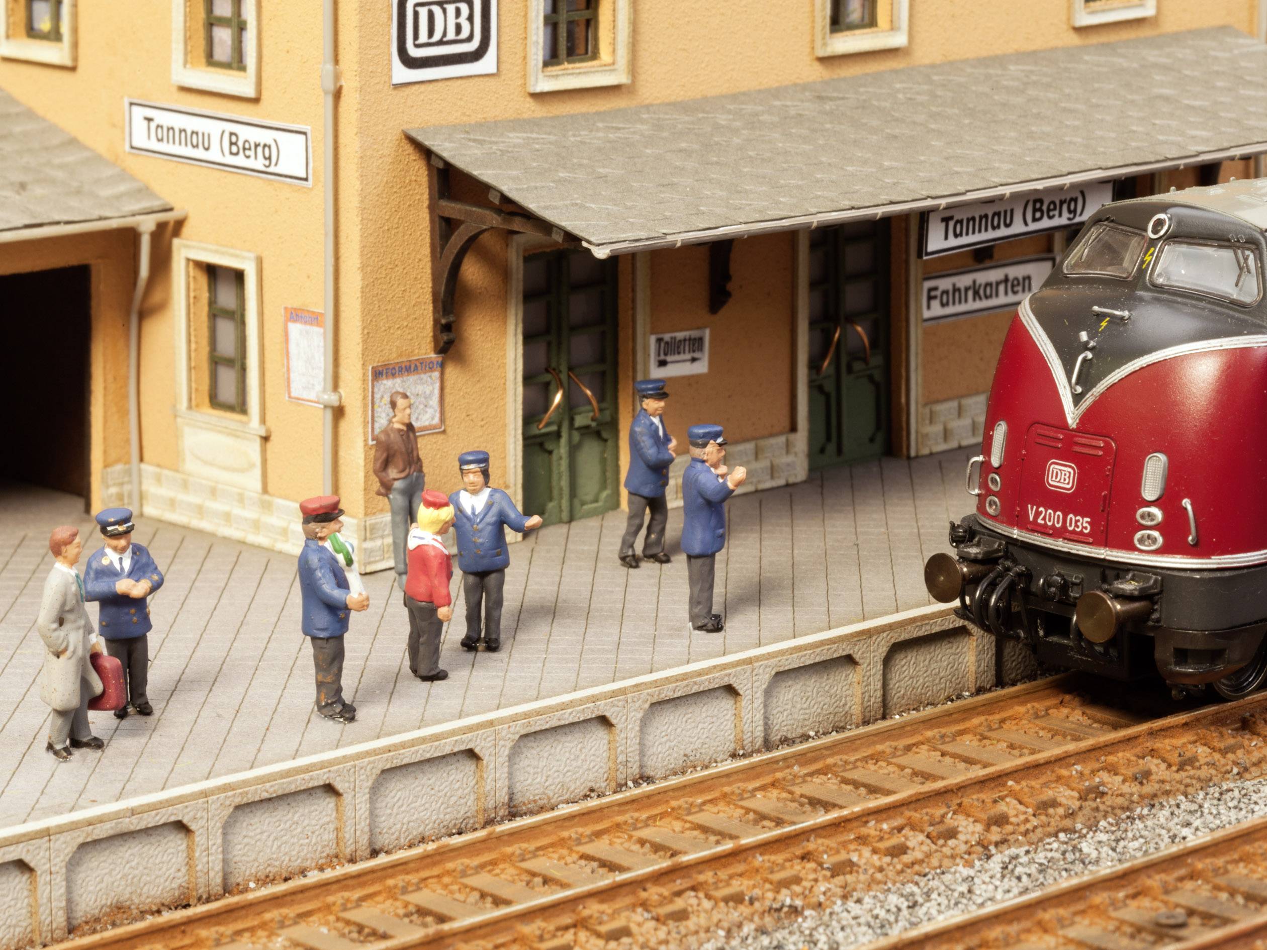 Une scène de train miniature montre une gare avec une locomotive rouge, des panneaux DB et des personnages en uniforme debout sur le quai.