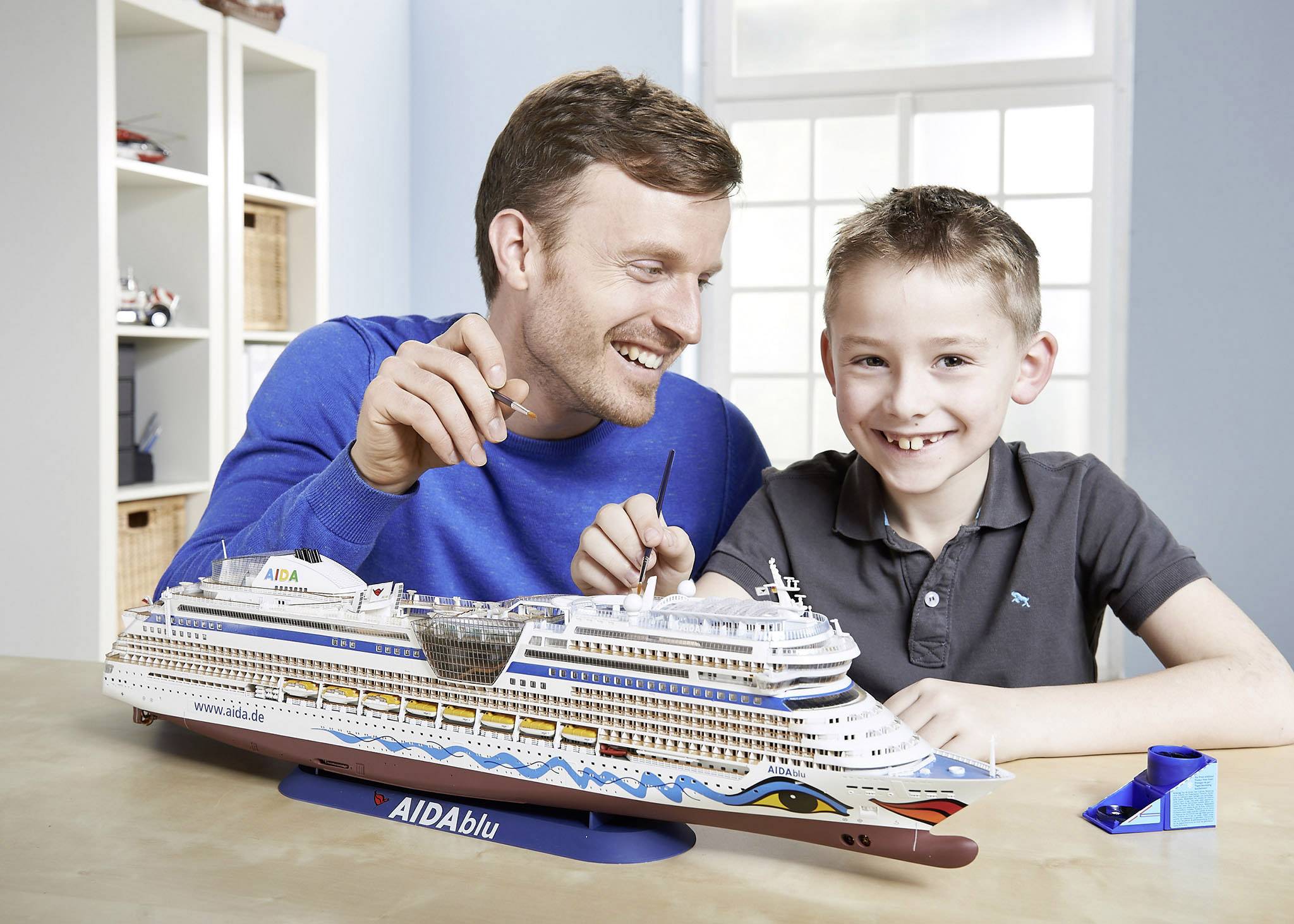 Un homme et un garçon construisent ensemble une maquette de bateau. Tous deux sourient et sont assis à une table avec le modèle du navire de croisière 'AIDABlu'.
