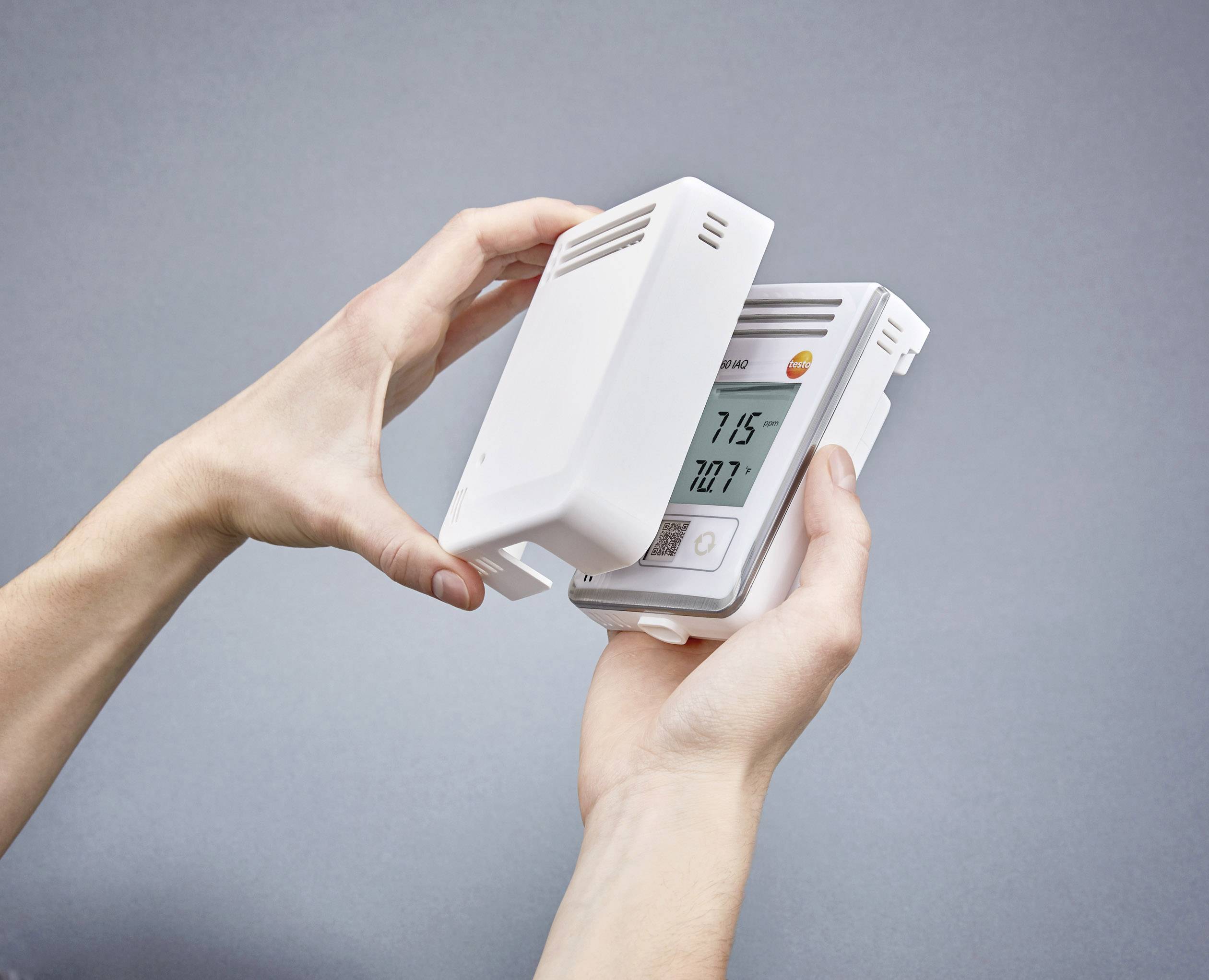 Une personne tient un thermostat dans ses mains. L'affichage indique une température de 21,5°C. L'appareil est dans un boîtier blanc.