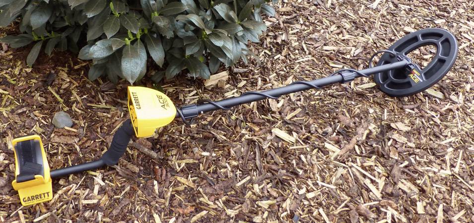 Un détecteur de métaux est posé sur des copeaux de bois, partiellement devant un buisson. L'appareil est noir et jaune, avec le nom de marque 'Garrett' bien visible.