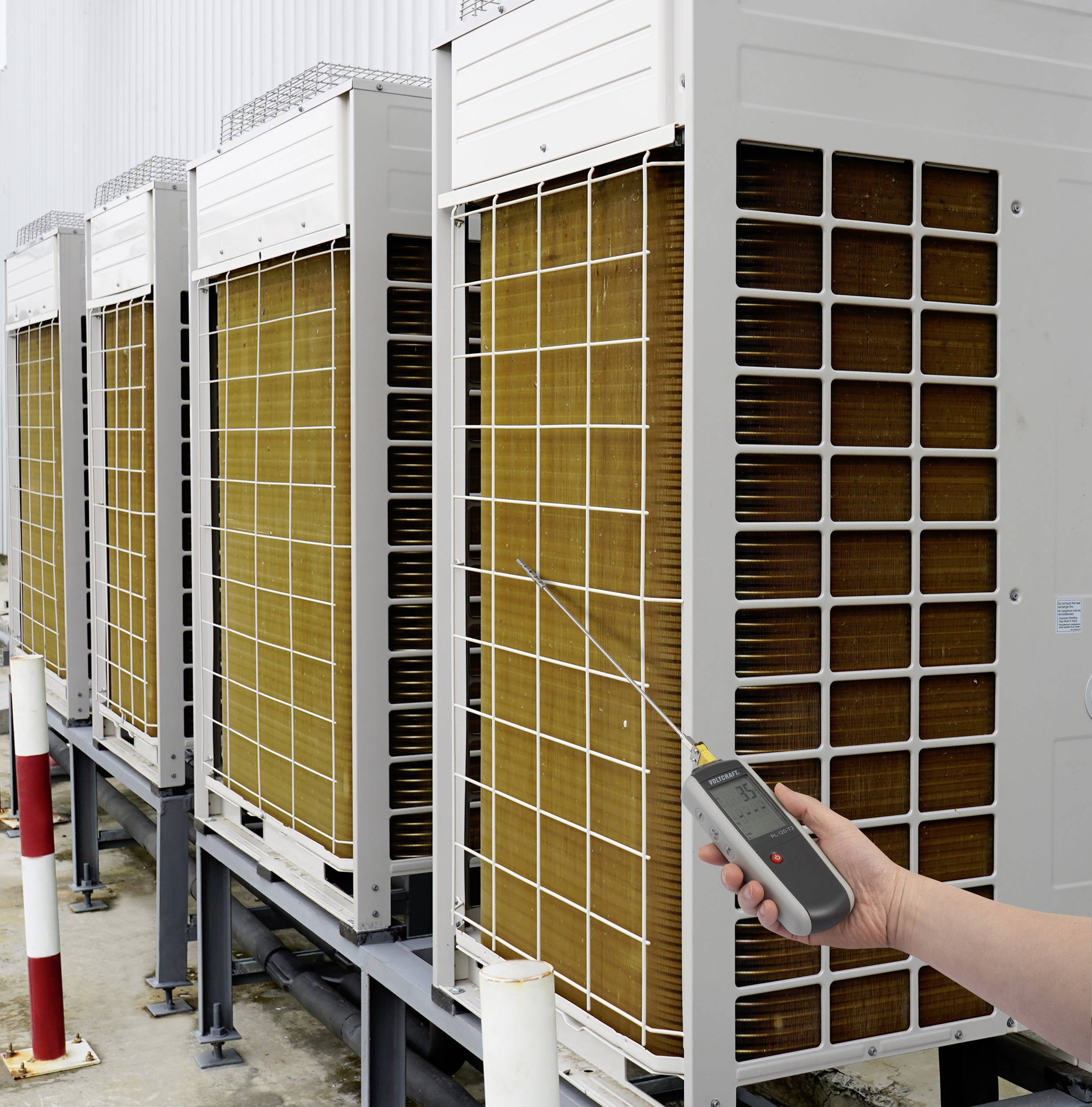 Une personne vérifie des unités extérieures de climatisation à l'aide d'un thermomètre numérique. Les appareils sont disposés en ligne.