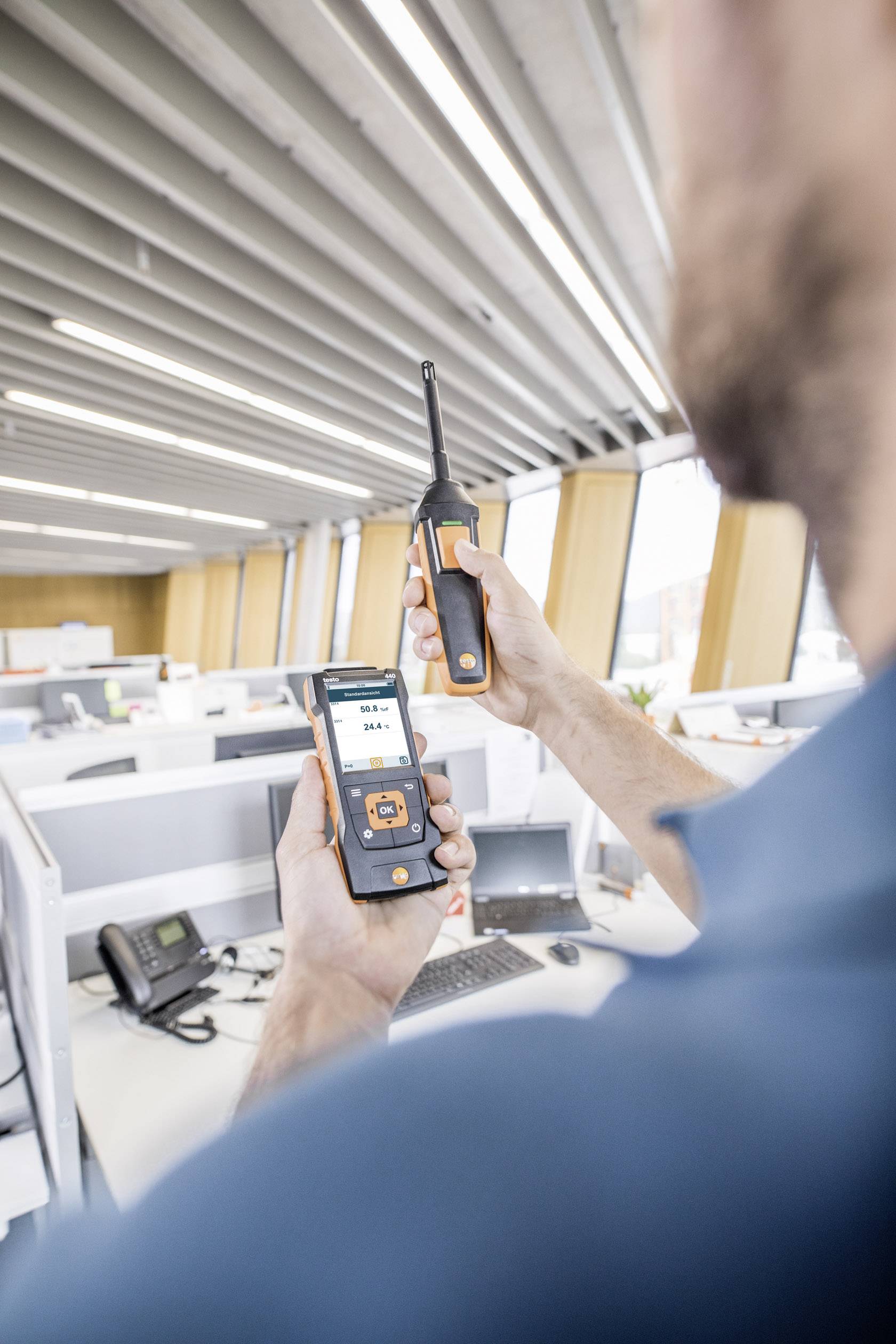Un homme dans un bureau utilise un appareil de mesure pour vérifier la qualité de l'air à un poste de travail. Plusieurs bureaux et ordinateurs sont visibles en arrière-plan.