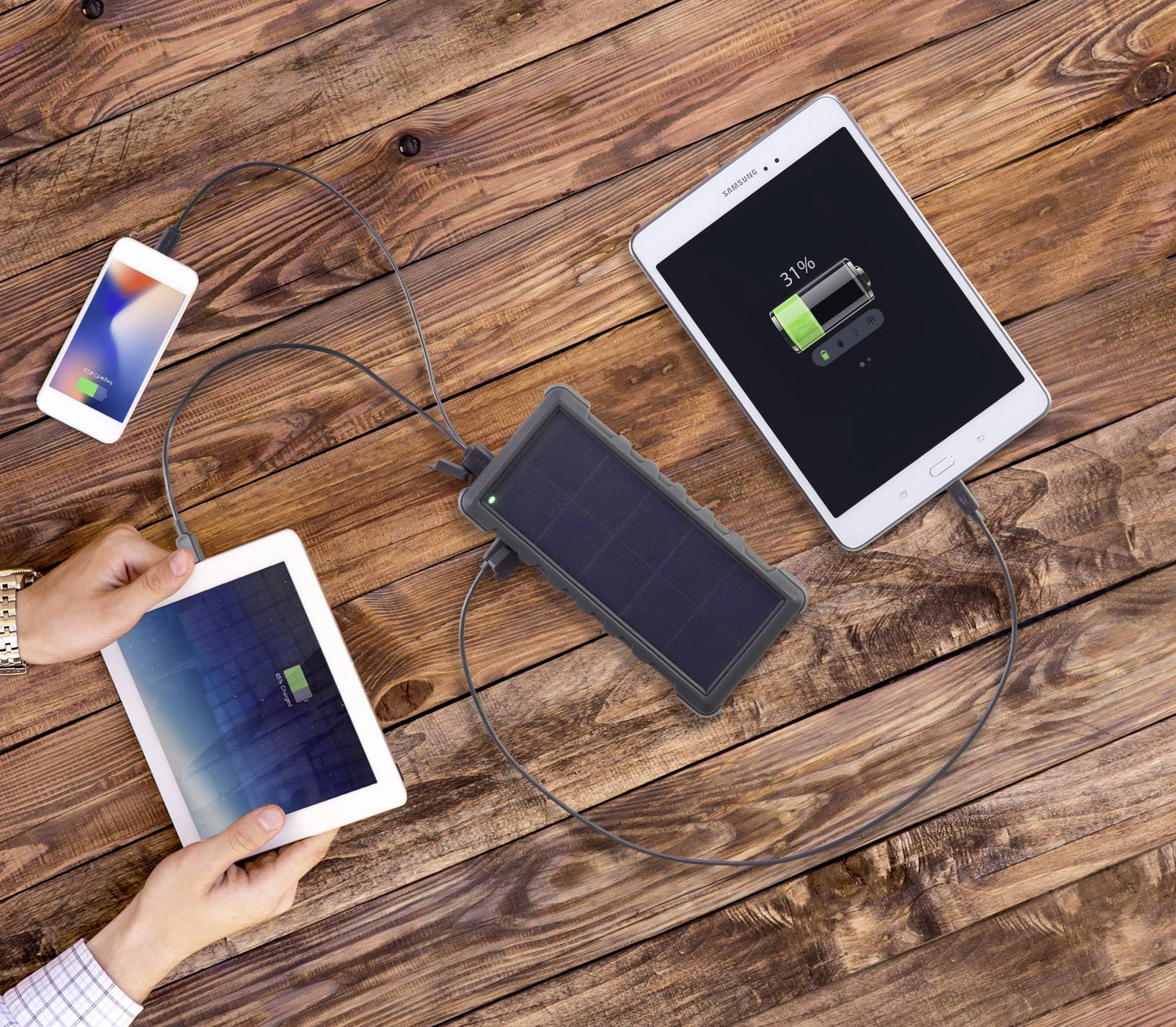 Un chargeur solaire posé sur une table en bois charge sans fil un smartphone, une tablette et un autre smartphone.