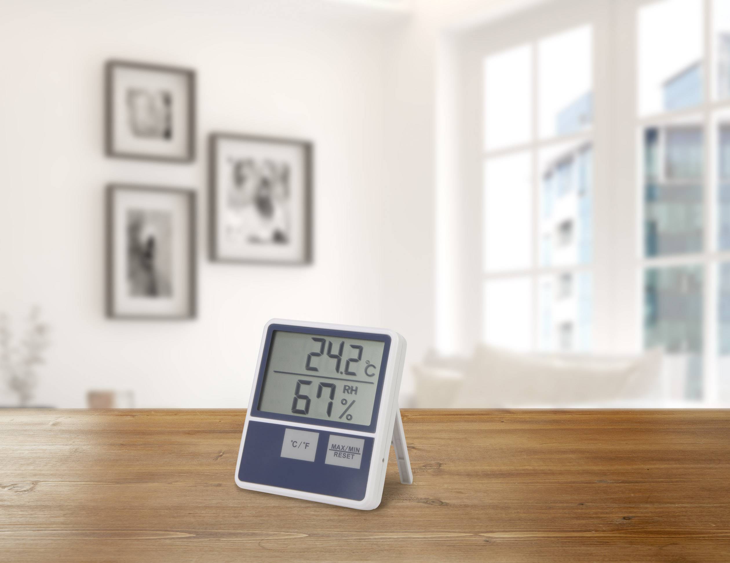 Un hygromètre numérique posé sur une table en bois affiche 24,2°C et 67% d'humidité relative. Des fenêtres et des images floues sont visibles en arrière-plan.