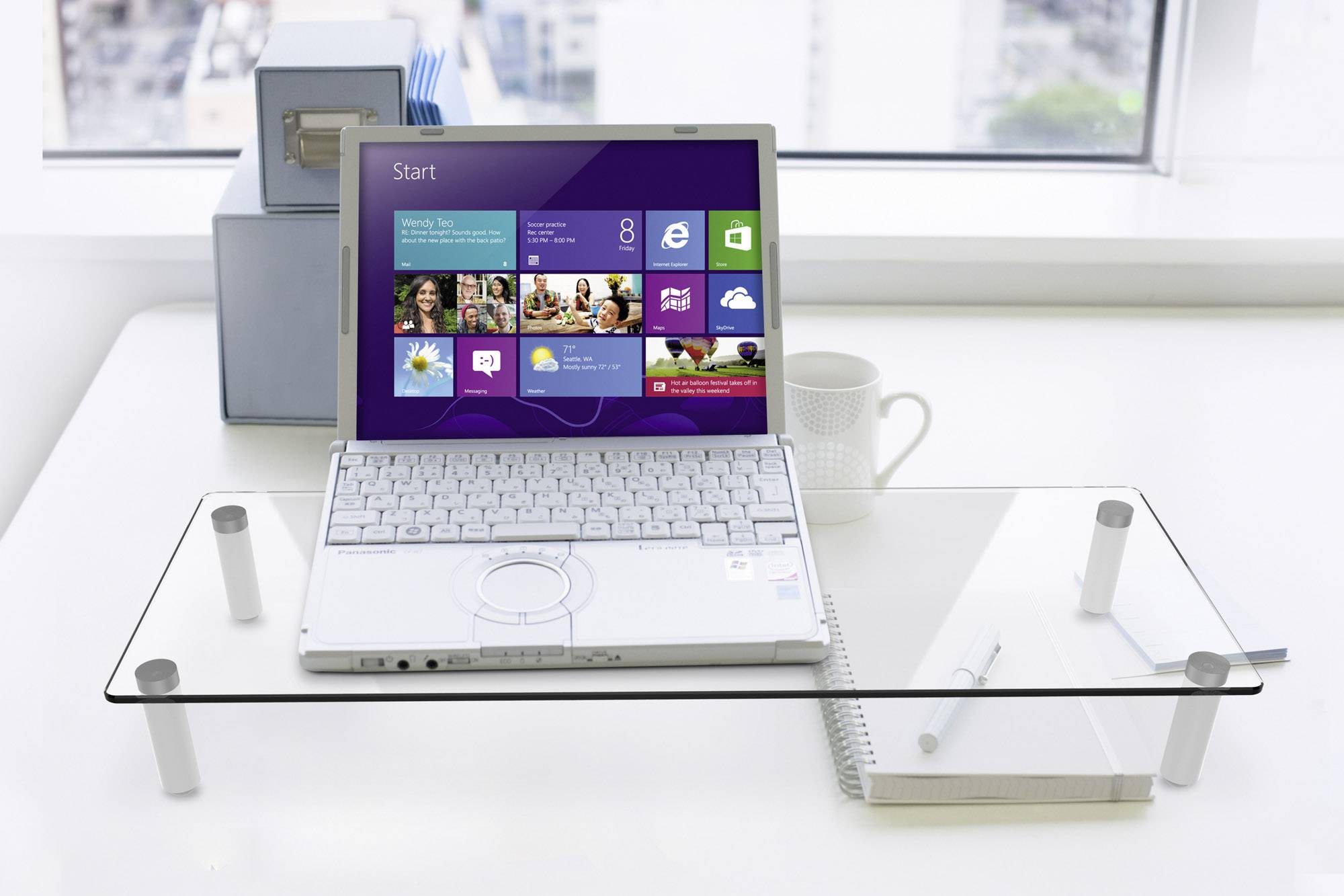 Un ordinateur portable sous Windows 8 est posé sur une table en verre. En arrière-plan, une vue par la fenêtre donne sur une ville. À côté se trouvent une tasse et un cahier.