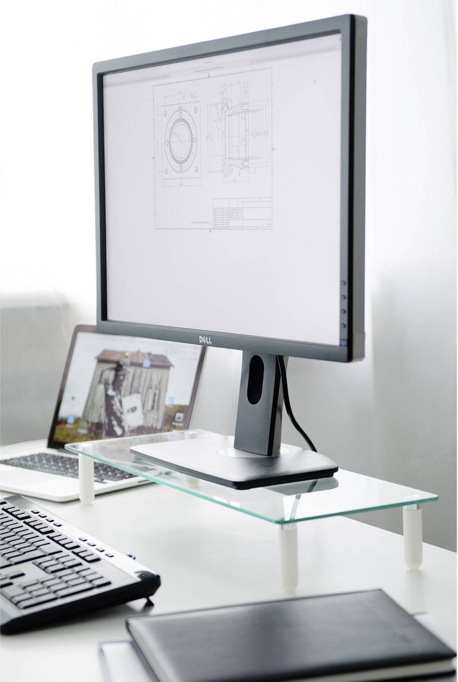 Un poste de travail informatique avec un grand écran affiche des dessins techniques. Un ordinateur portable est en arrière-plan. Un clavier et un carnet de notes sont posés sur la table.