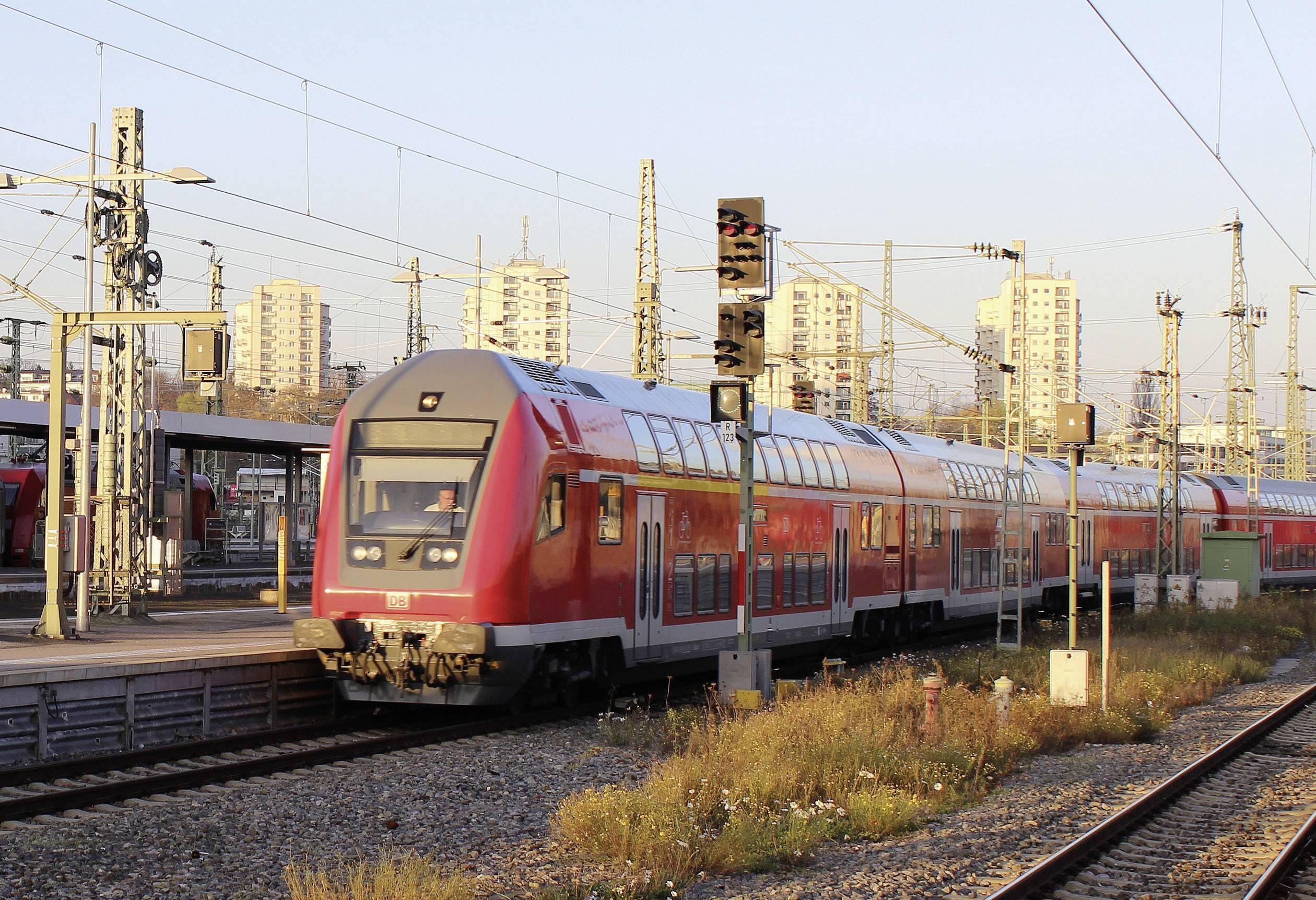 Un train de transport urbain rouge entre en gare pendant la journée. En arrière-plan, on peut voir des immeubles de grande hauteur et des pylônes électriques.