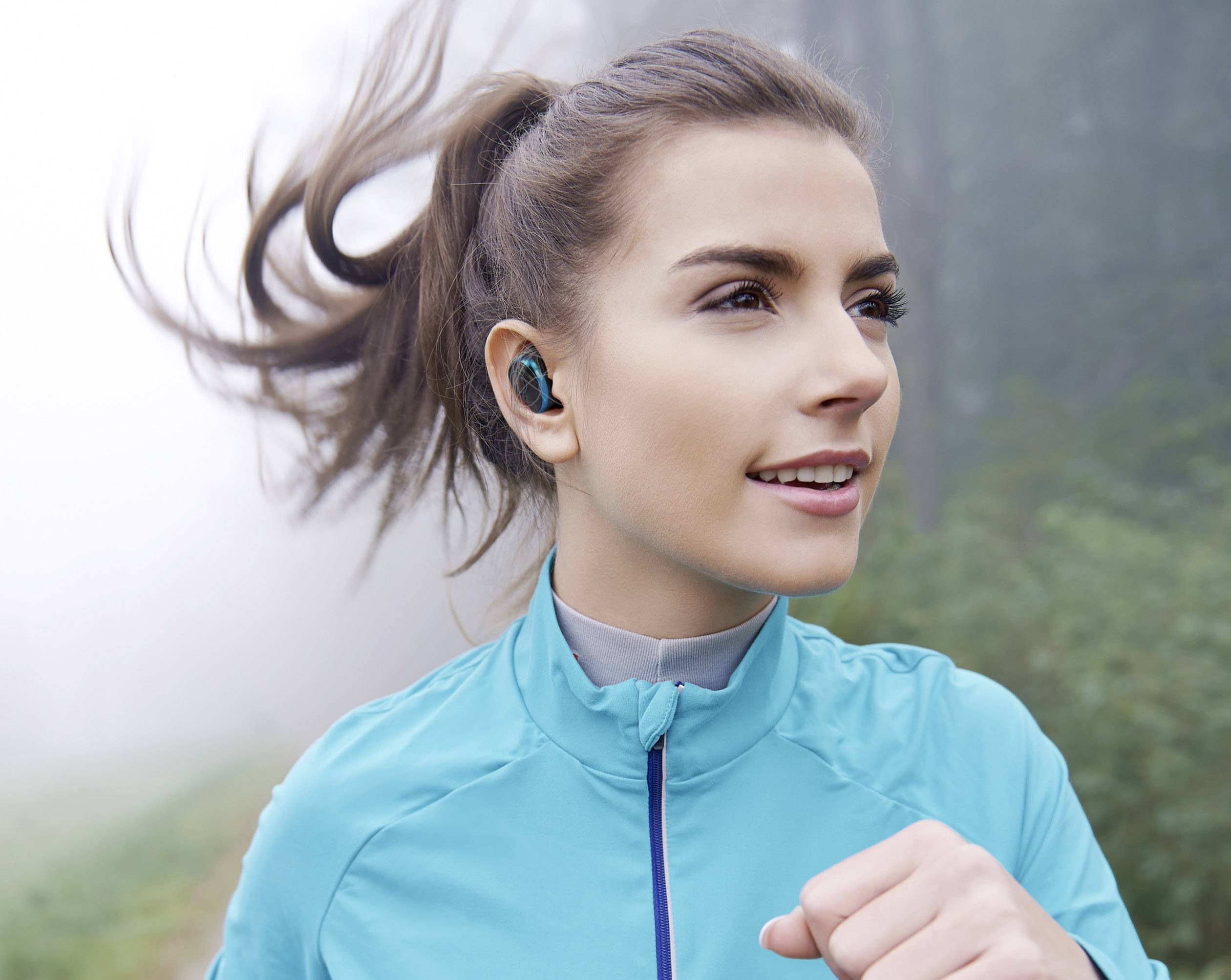 Une femme fait du jogging portant une veste bleue par un jour de brume. Elle a des écouteurs sans fil et regarde devant elle avec concentration.