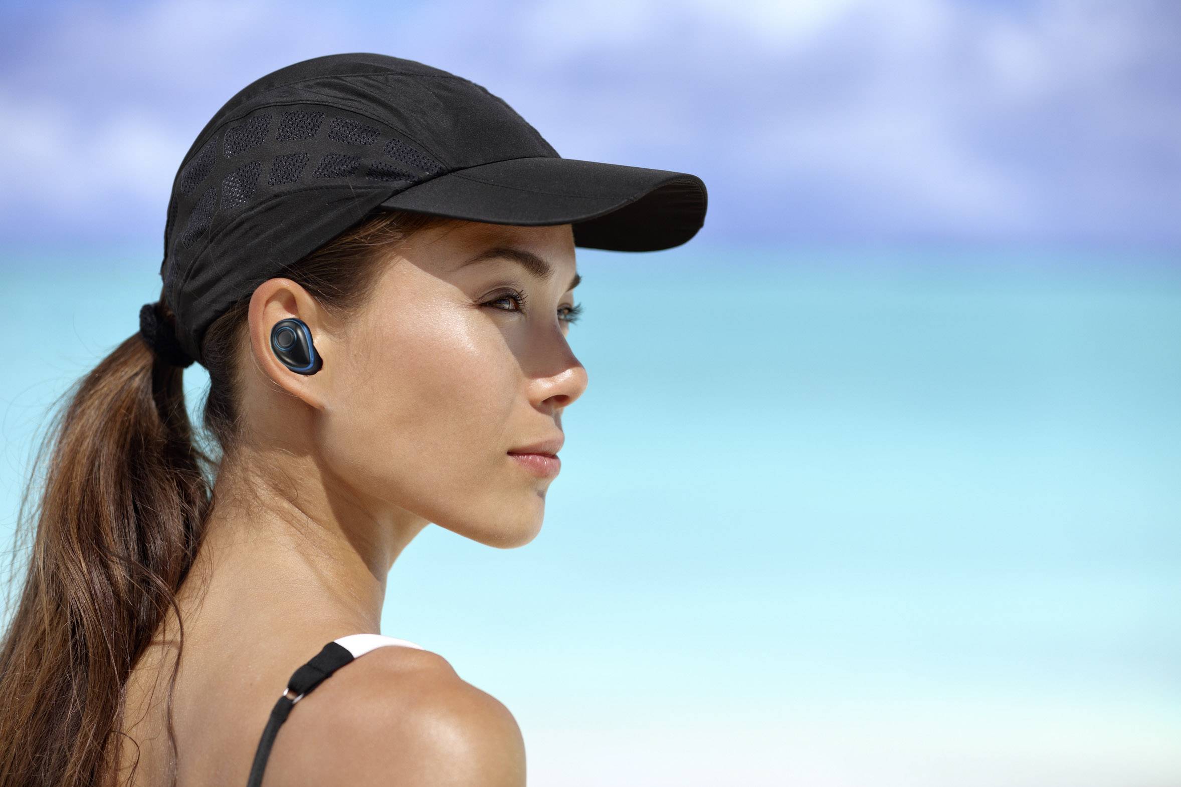 Femme sur la plage portant une casquette noire et des écouteurs sans fil, regardant la mer. L'arrière-plan présente une eau et un ciel bleu clair.