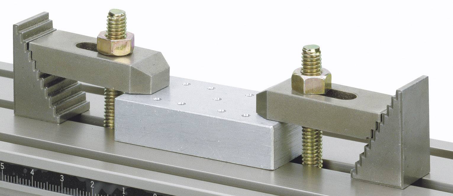 Un étau maintient une pièce rectangulaire de métal. Deux supports munis de vis fixent la pièce sur un rail de guidage.