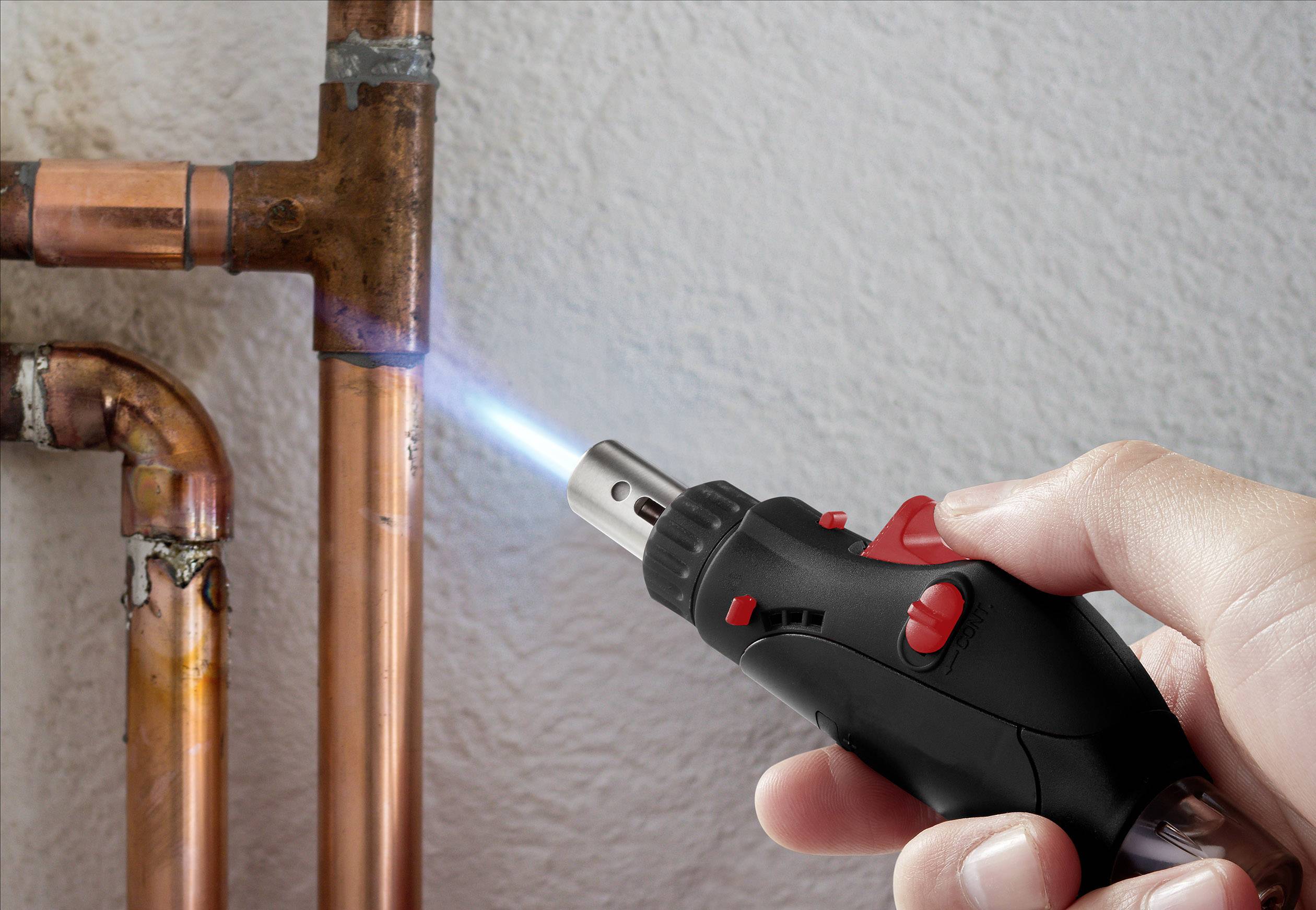 Une main tient un petit brûleur et chauffe un tube de cuivre pour souder une connexion.