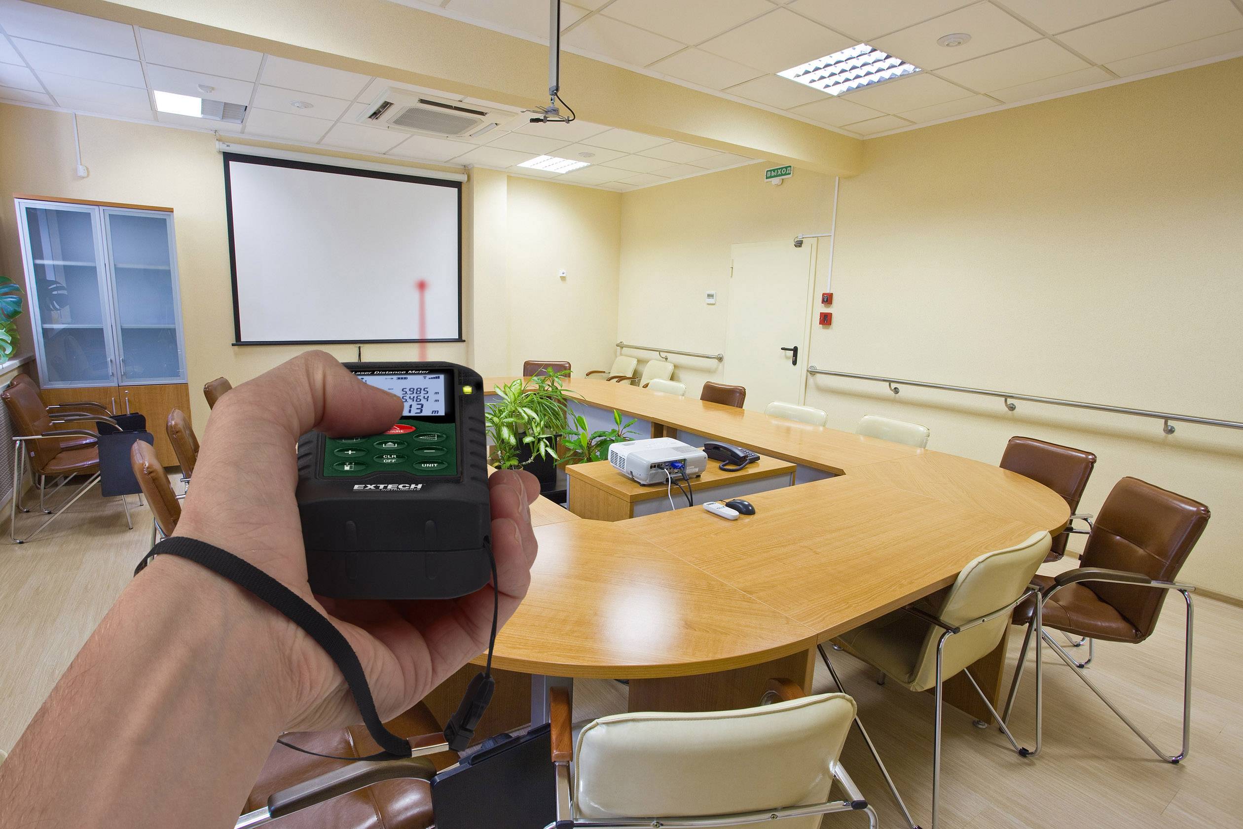 Une personne tient un télémètre dans une salle de conférence. Un point laser rouge atteint l'écran. Dans la pièce, il y a une table ovale avec des chaises.