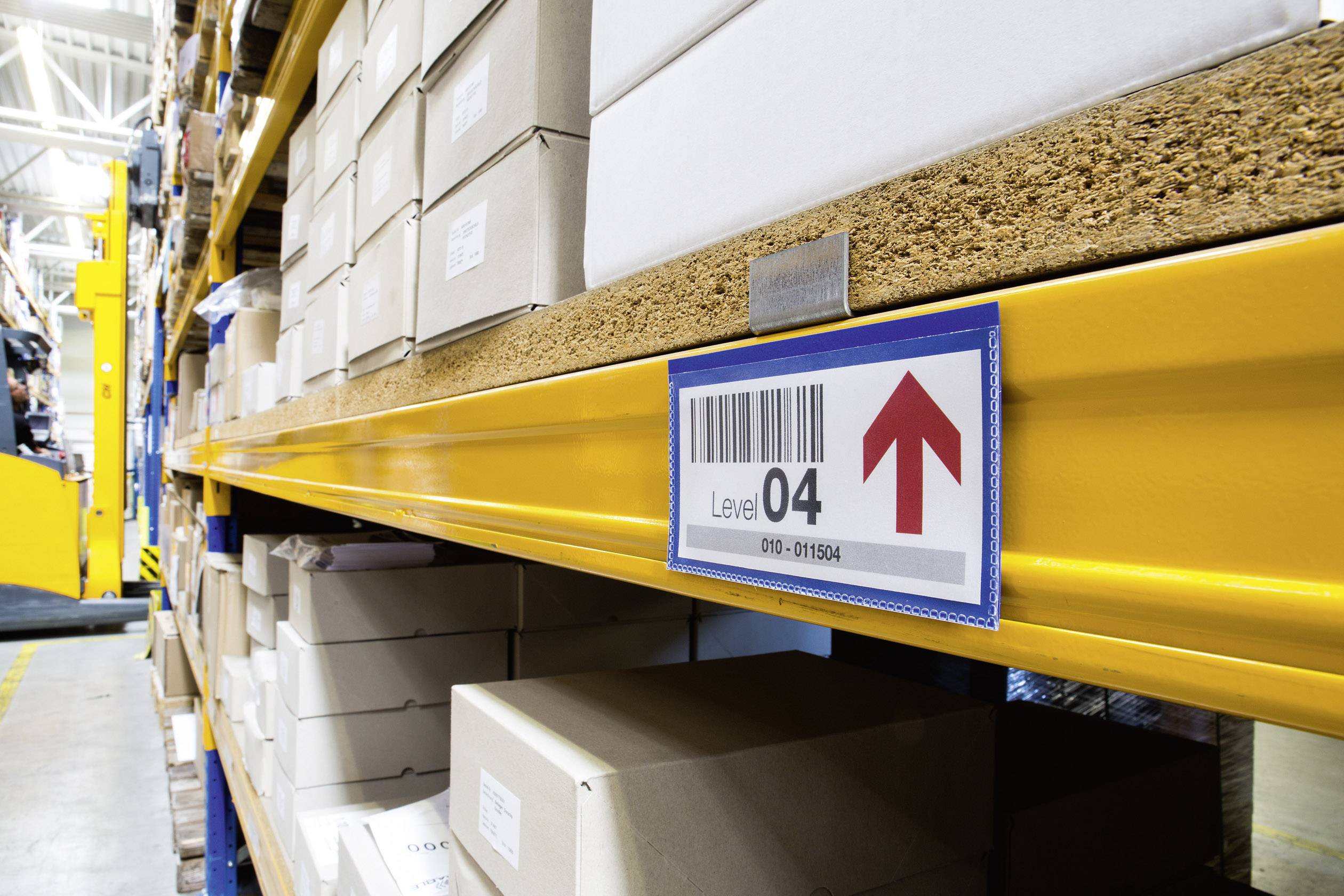 Rangée d'étagères dans un entrepôt avec des boîtes empilées. Un panneau avec un code-barres pointe vers le haut et indique le niveau 4. Un chariot élévateur est visible en arrière-plan.
