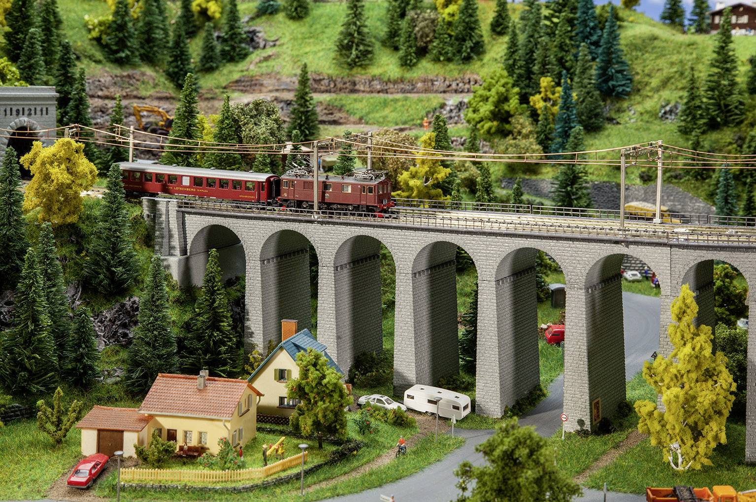 Un train rouge circule sur un haut viaduc dans un paysage de modélisme rural avec des arbres, des routes et de petites maisons.