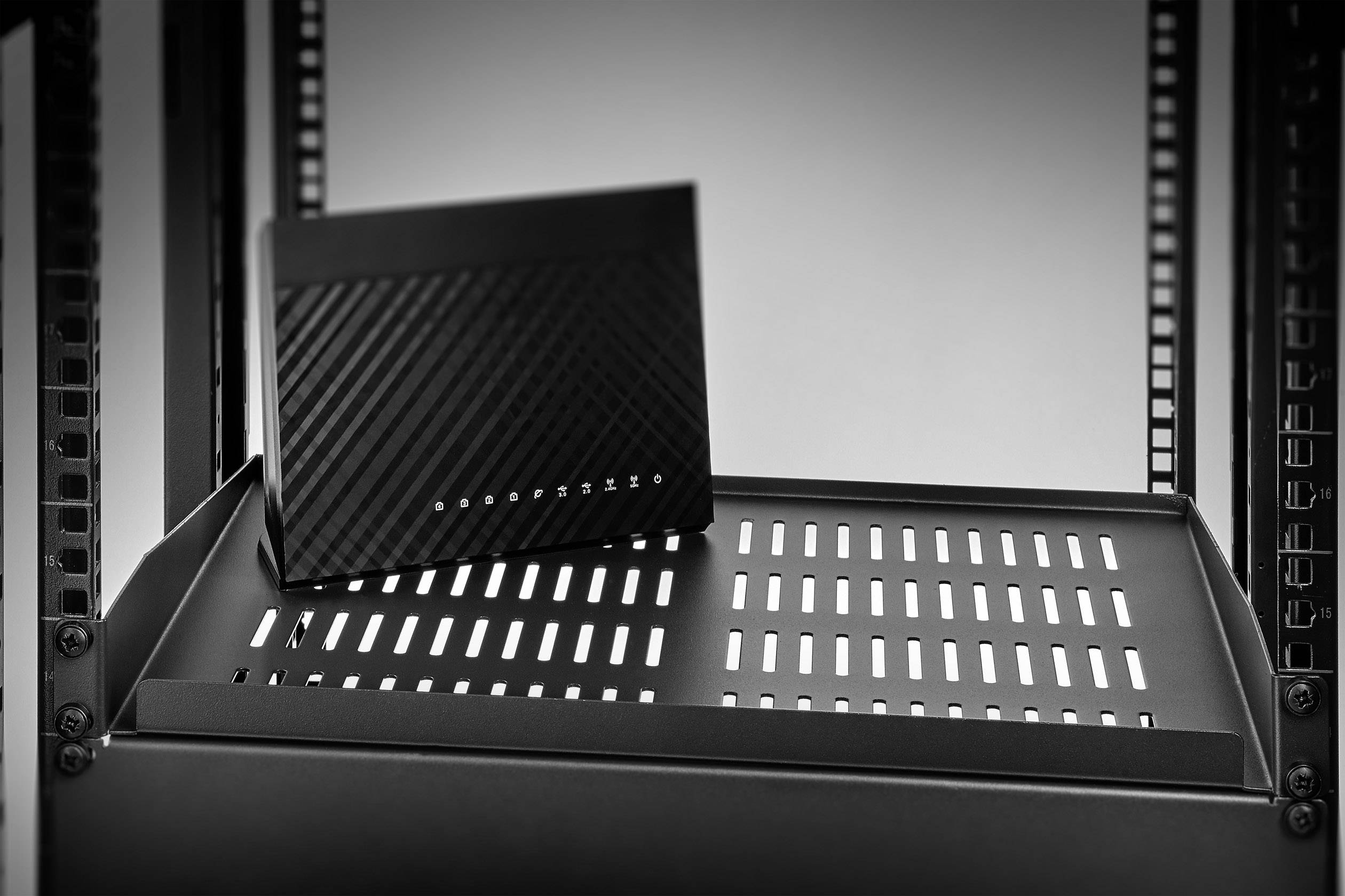 Un routeur noir avec des fentes de ventilation est posé sur une étagère dans un rack serveur, sur un fond neutre.