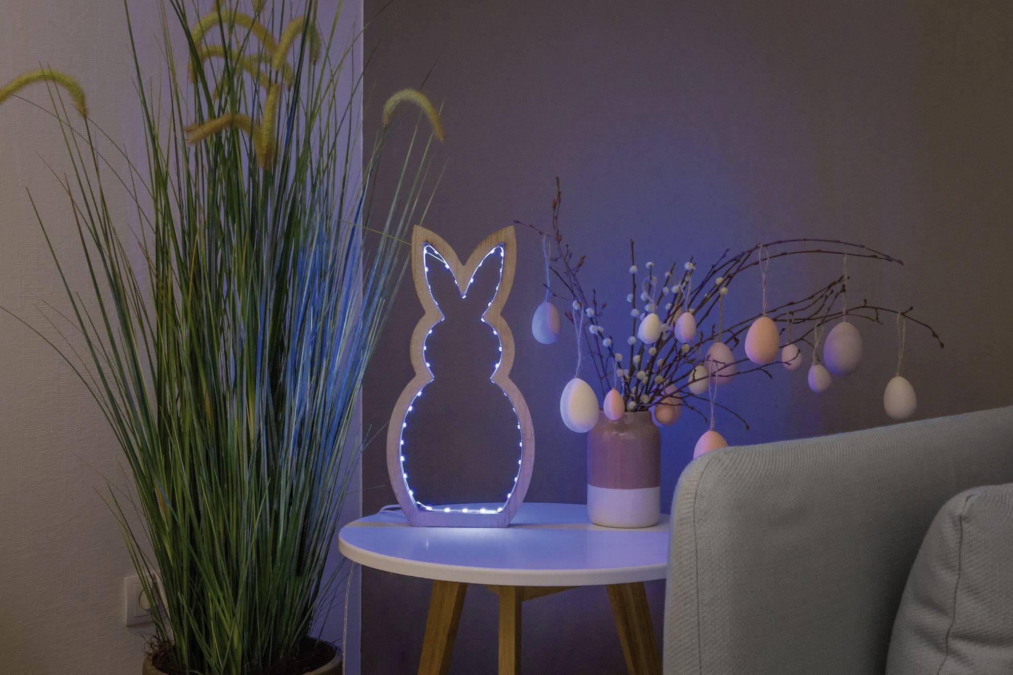 Table décorée pour Pâques : Cadre lumineux en forme de lapin et branches de Pâques avec des œufs peints, entourés de plantes d'intérieur et de mobilier moderne.