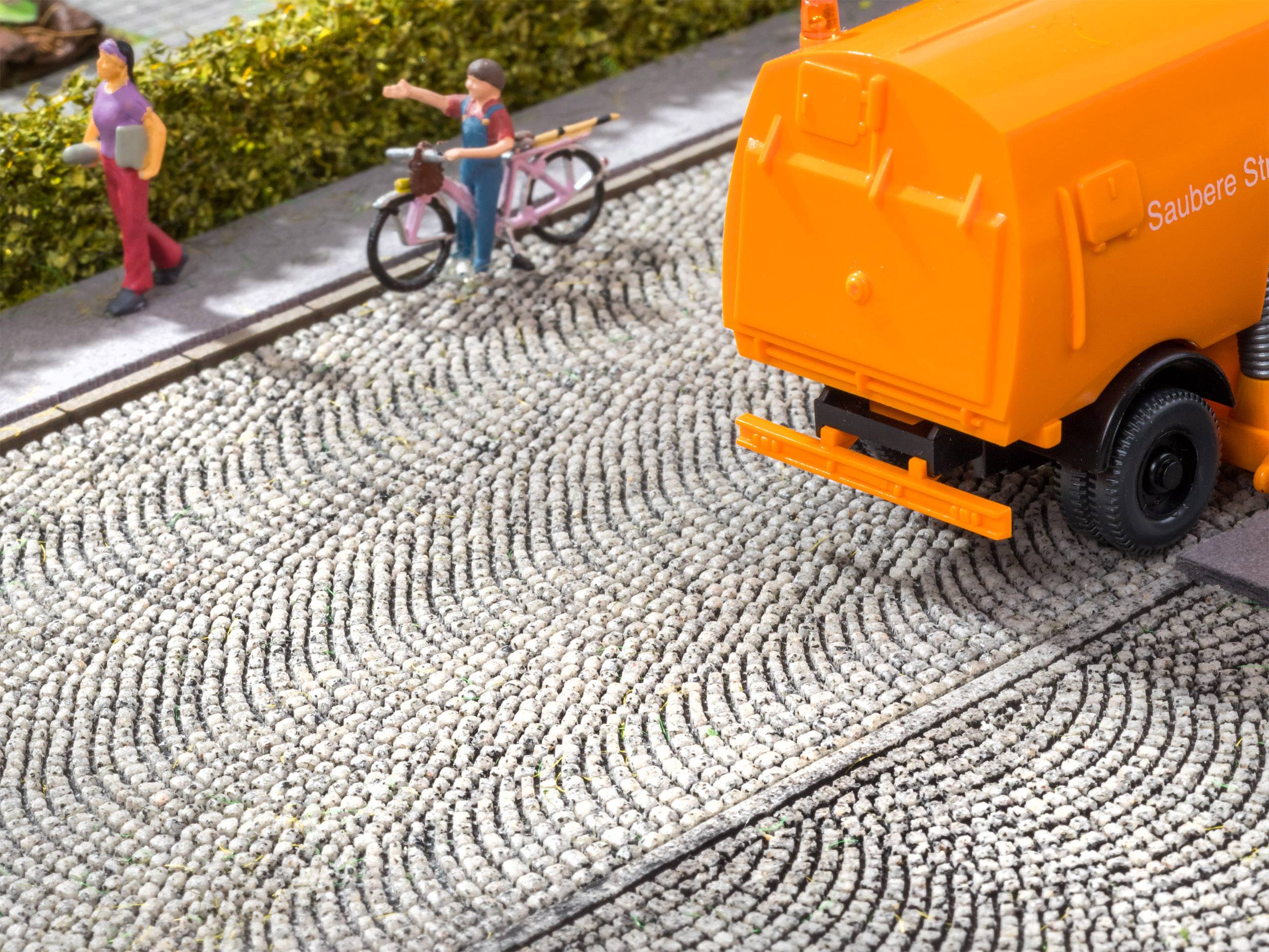 Un petit modèle représente un balai de rue orange sur une route pavée, avec deux personnages et un vélo en bordure.