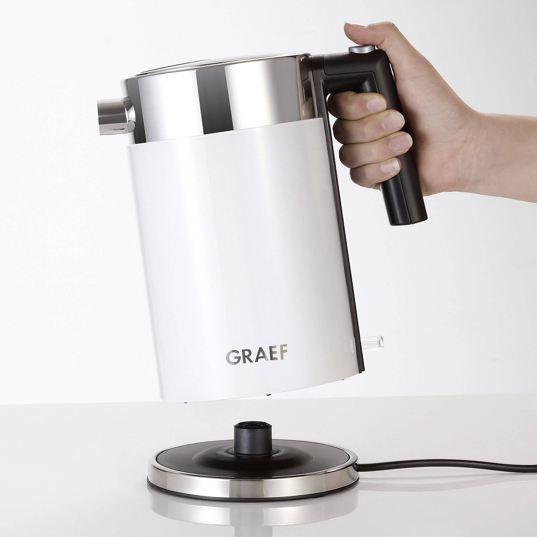 Bouilloire électrique au design moderne posée sur sa base, tenue par une main par la poignée. Nom de marque 'GRAEF' visible sur l'avant.