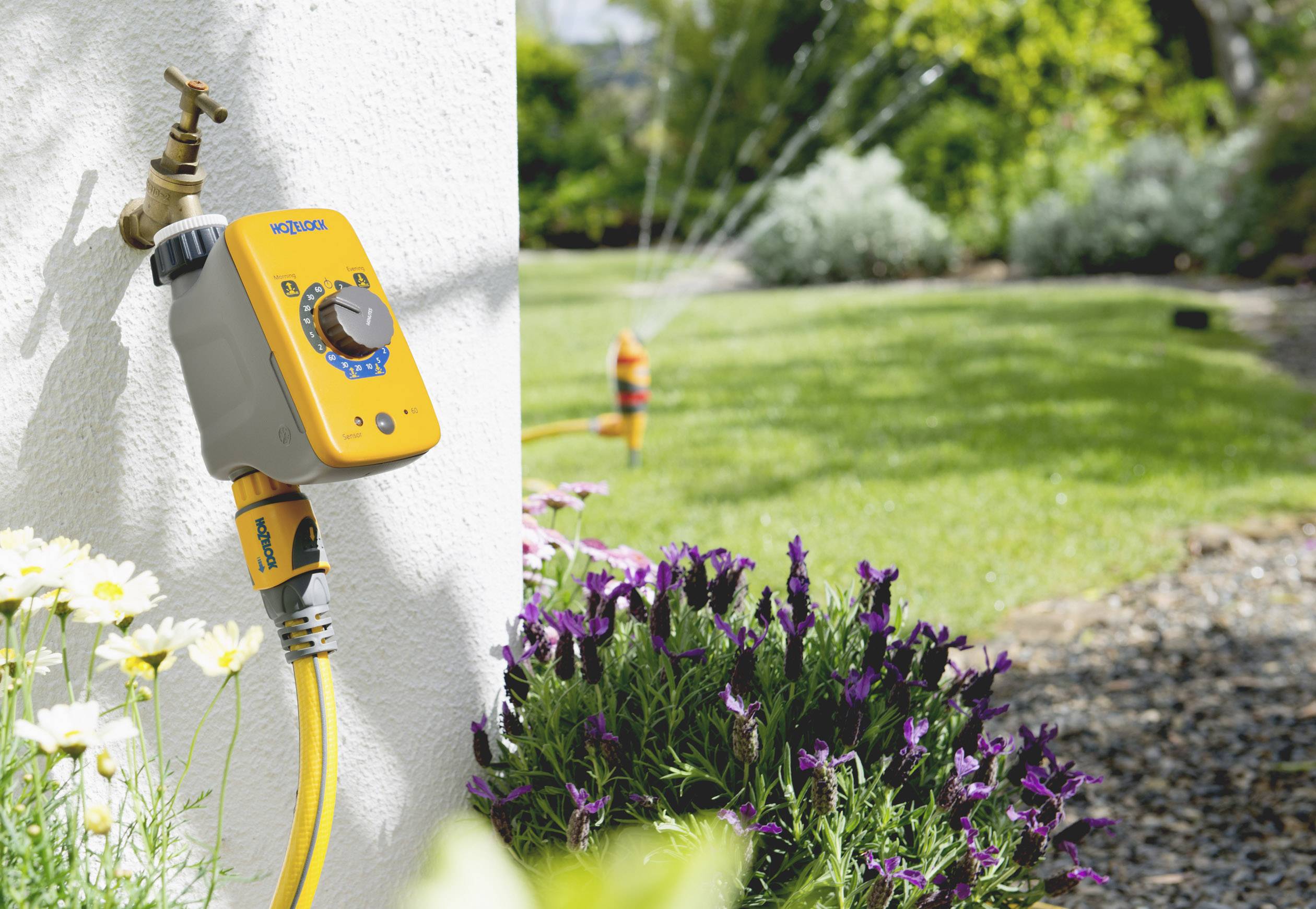 Système d'irrigation de jardin avec un minuteur automatique installé sur un robinet. En arrière-plan, des arroseurs aspergent un gazon vert.