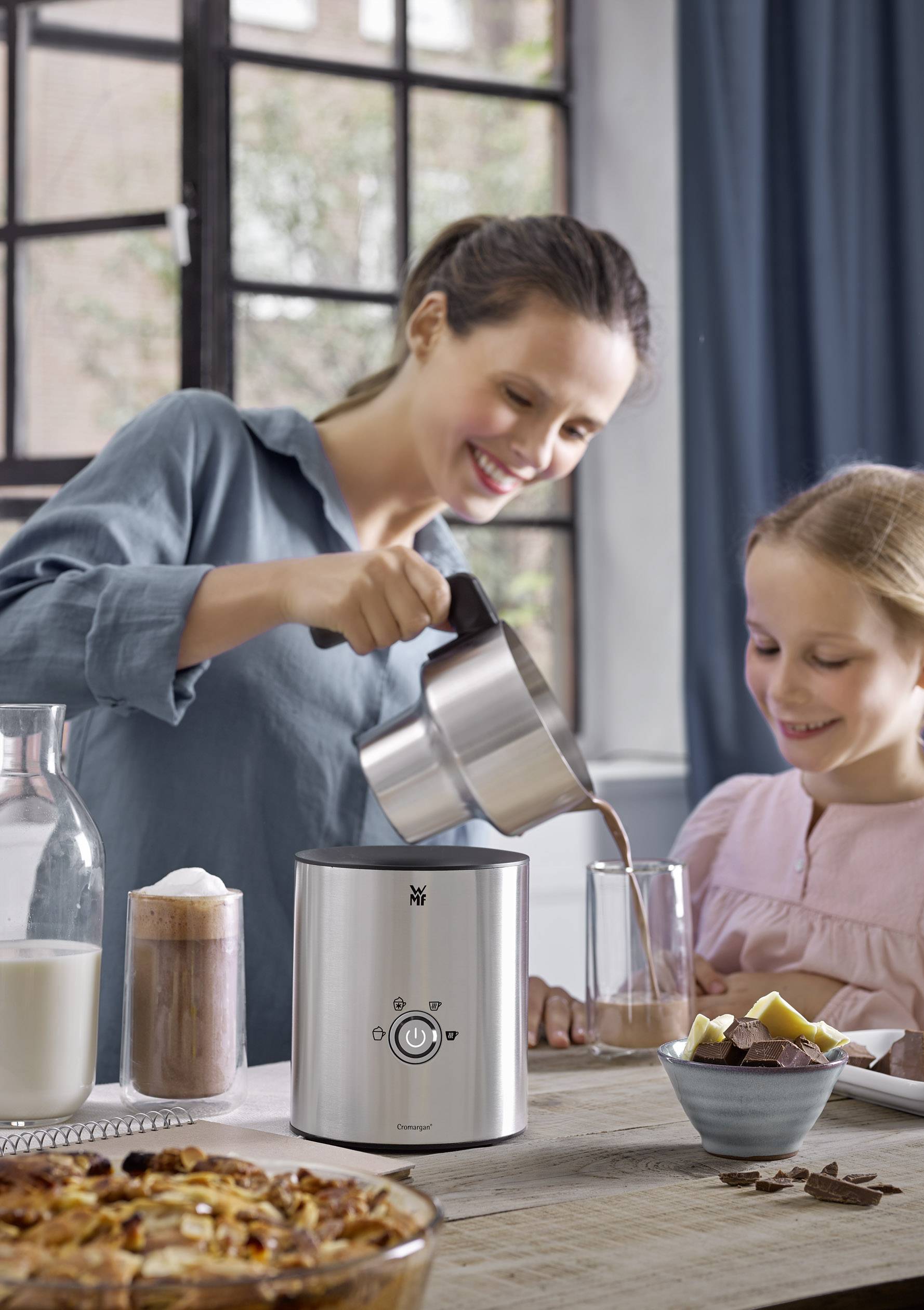 Une femme verse une boisson chaude dans un verre tandis qu'une jeune fille se tient à côté d'elle et sourit. Sur la table se trouvent du lait, des boissons et des collations.