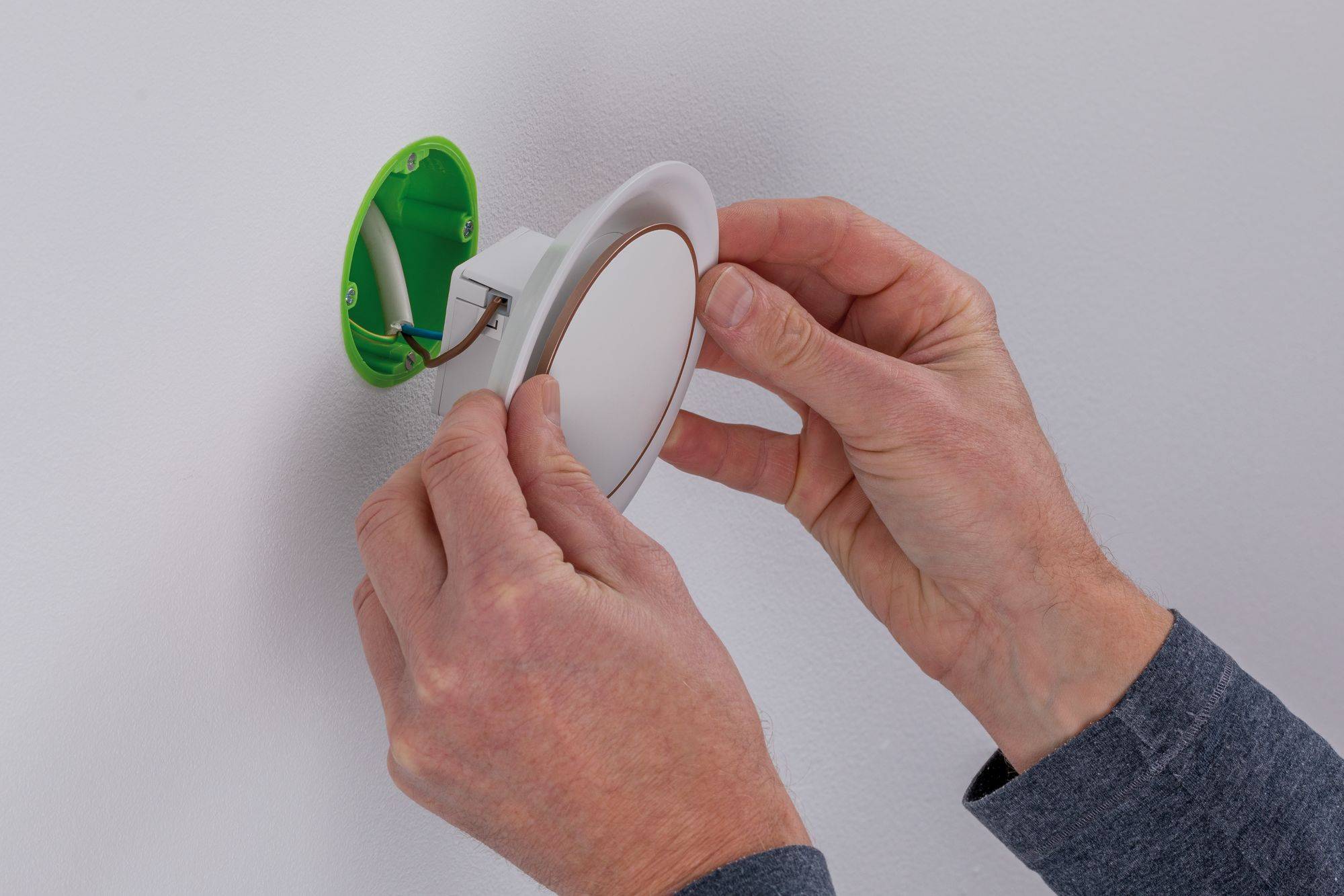 Une personne installe une lampe LED ronde dans une prise électrique verte ouverte sur un mur blanc.