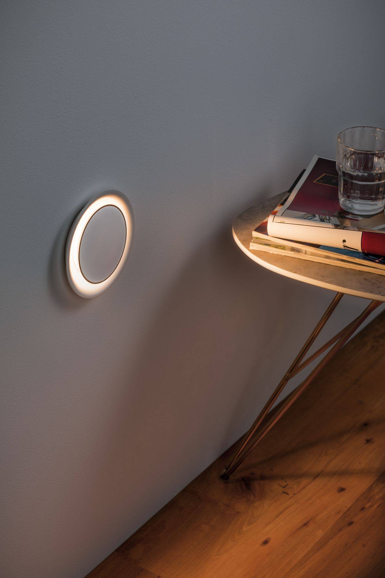 Une lampe ronde éclaire le mur. À côté, une table à trois pieds est posée sur un sol en bois, portant des livres et un verre d'eau.