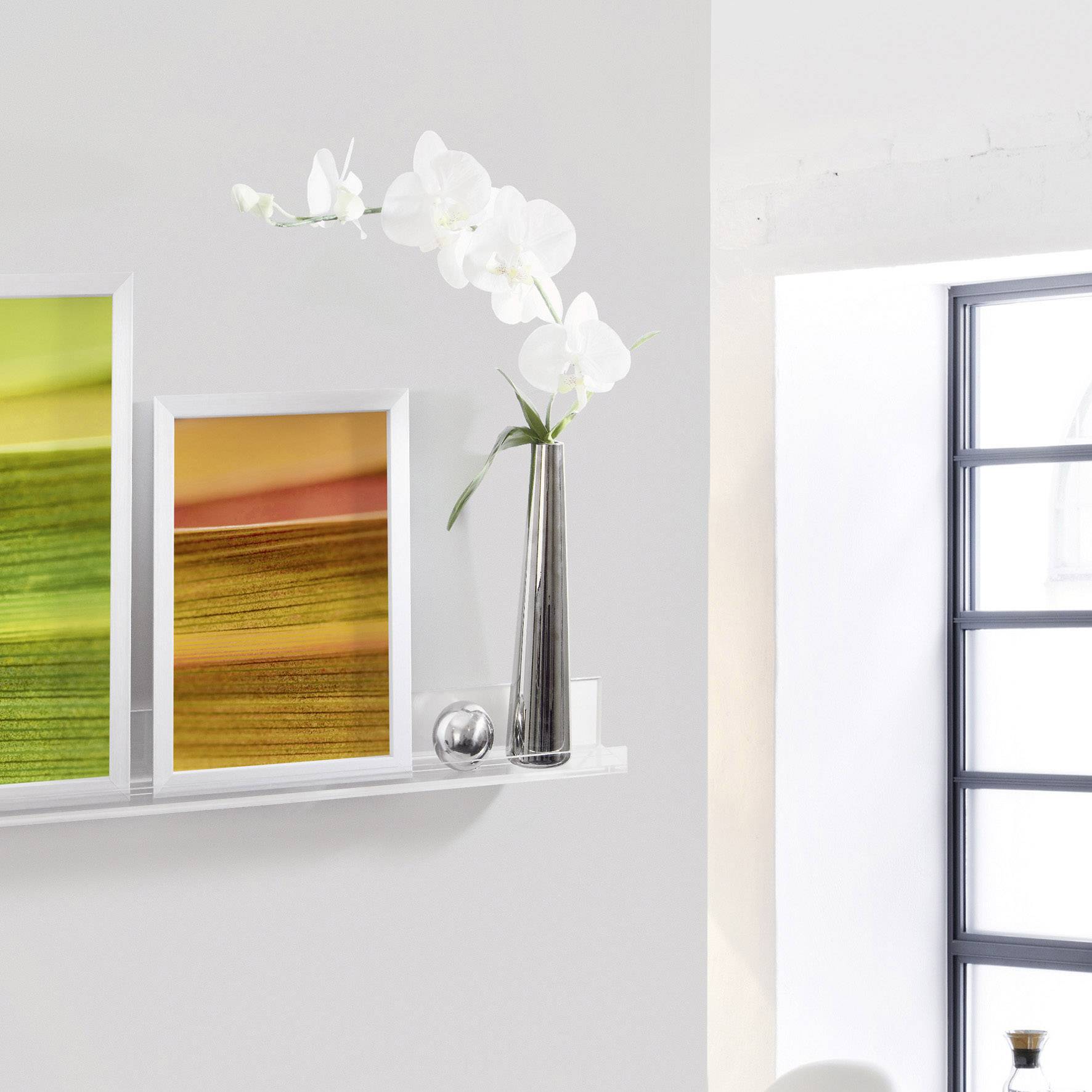 Murs blancs avec une étagère : Deux tableaux colorés et un vase contenant des orchidées blanches. Une lumière claire provenant de la fenêtre baigne la scène.