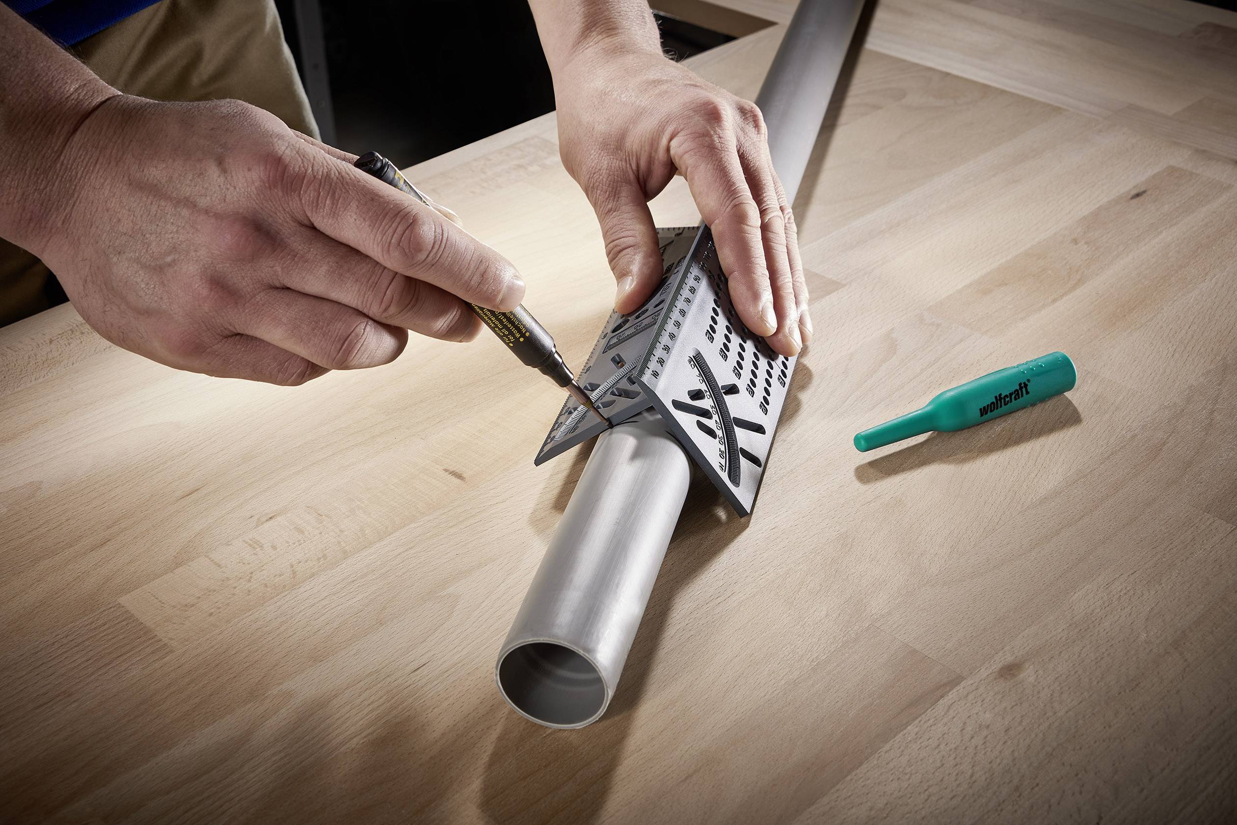 Une personne marque des mesures sur un tube métallique à l'aide d'un mètre et d'un stylo, alors que le tube est posé sur une table en bois. Un stylo vert est à proximité.