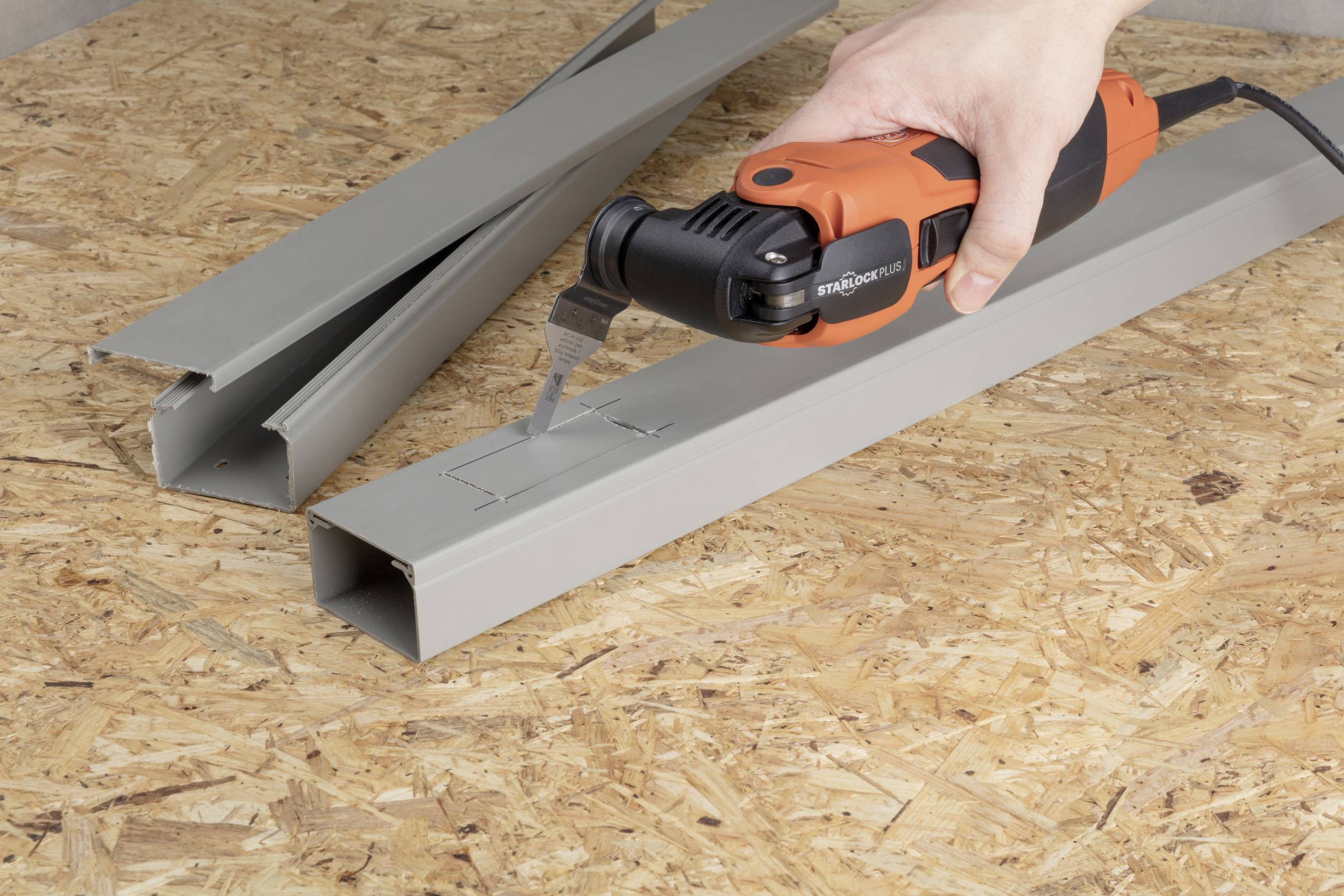 Une main tient un outil multifonction qui coupe un rectangle dans un profilé métallique. Le profilé est posé sur une surface en bois.