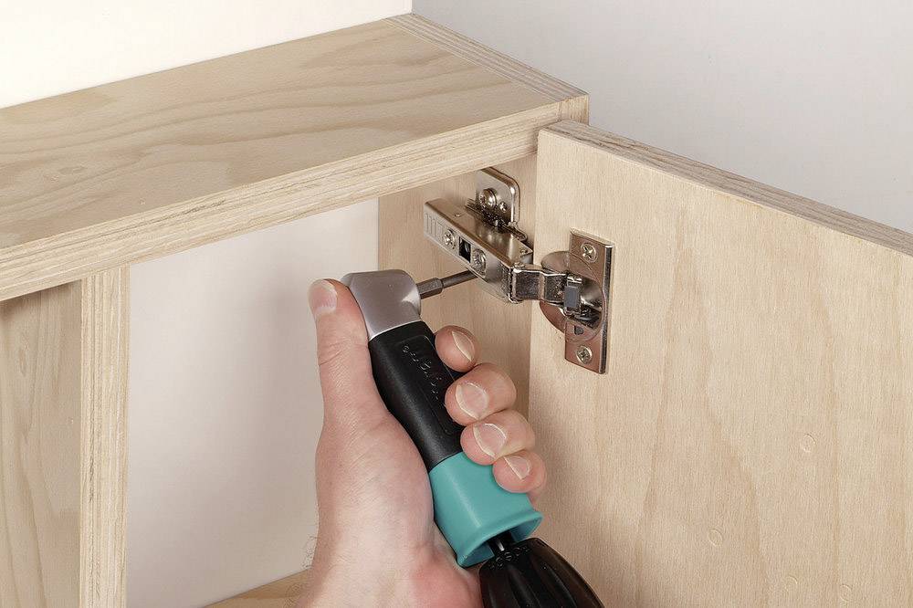 Une main visse une charnière de porte sur une armoire en bois à l'aide d'un tournevis.