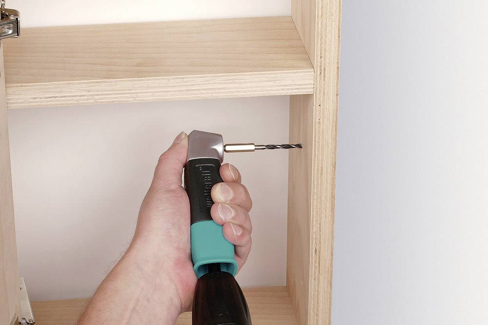 Une main tient une perceuse avec un adaptateur d'angle, en train de percer une étagère en bois. L'adaptateur permet de percer dans des espaces restreints.