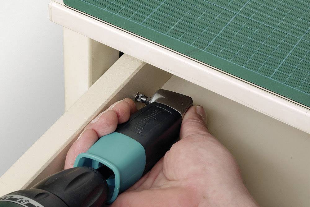 Une main utilise une visseuse sans fil pour serrer une vis dans un tiroir. En haut à gauche se trouve un tapis de coupe vert.