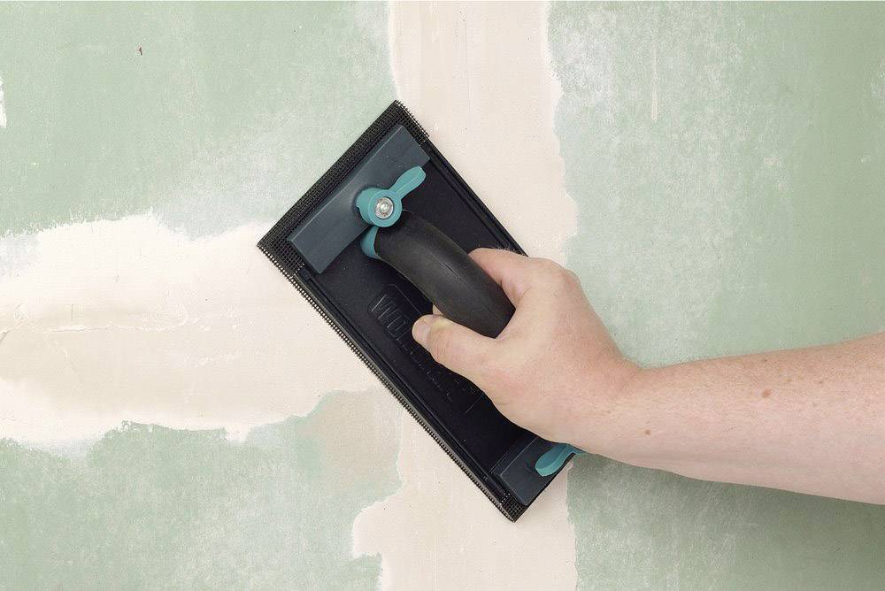 Une main lisse un mur enduit à l'aide d'un bloc de ponçage. L'arrière-plan montre une surface aux joints travaillés.