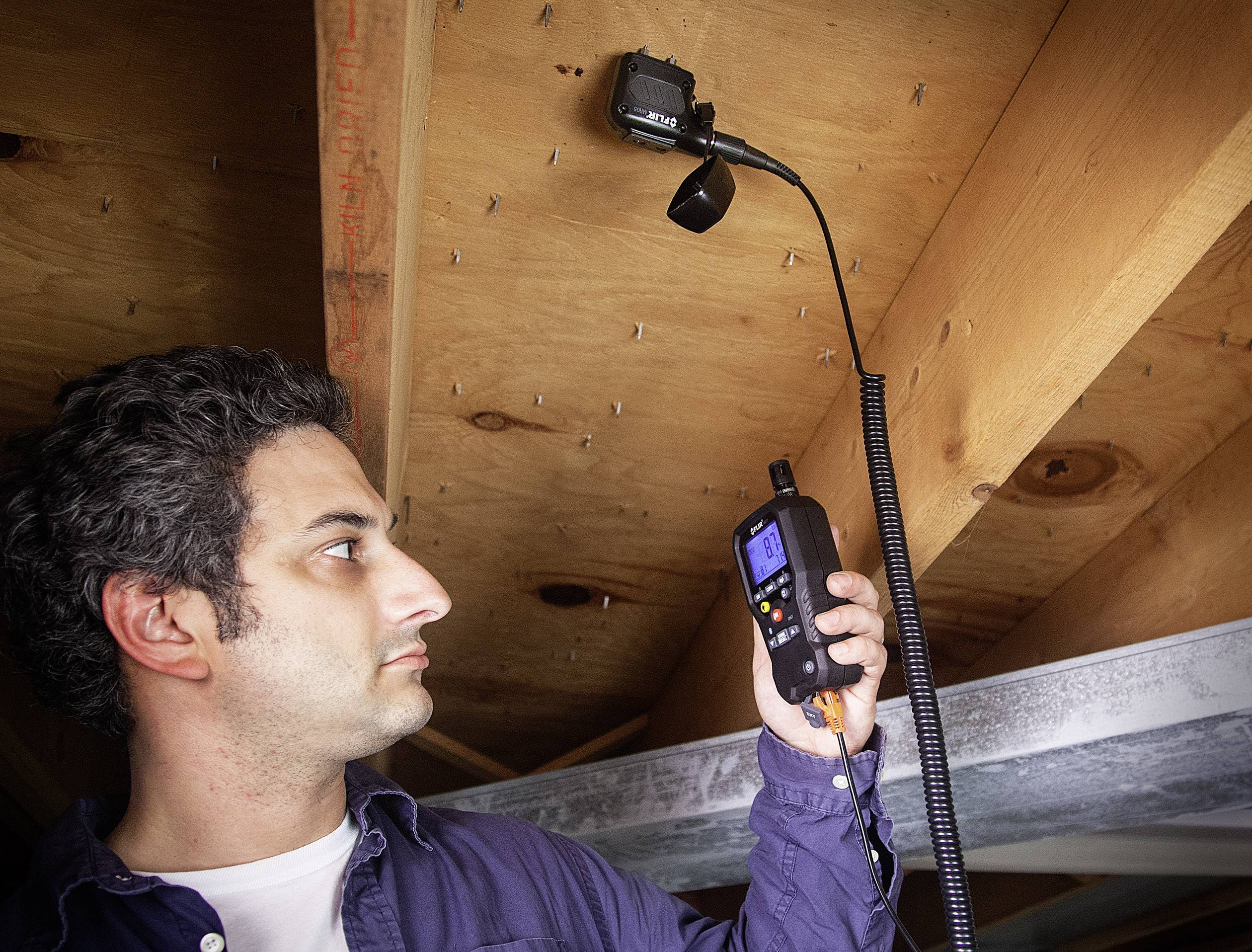 Un homme utilise un dispositif de caméra d'inspection pour examiner l'espace entre les poutres en bois. La caméra est flexible et dispose d'un écran.