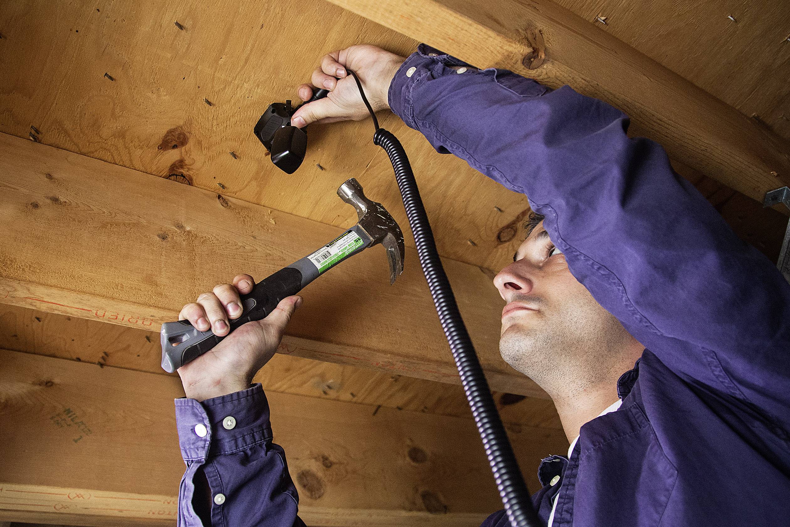 Un homme vêtu de bleu fixe une caméra au plafond en bois d'un bâtiment à l'aide d'un marteau. Il maintient la caméra avec son autre main.