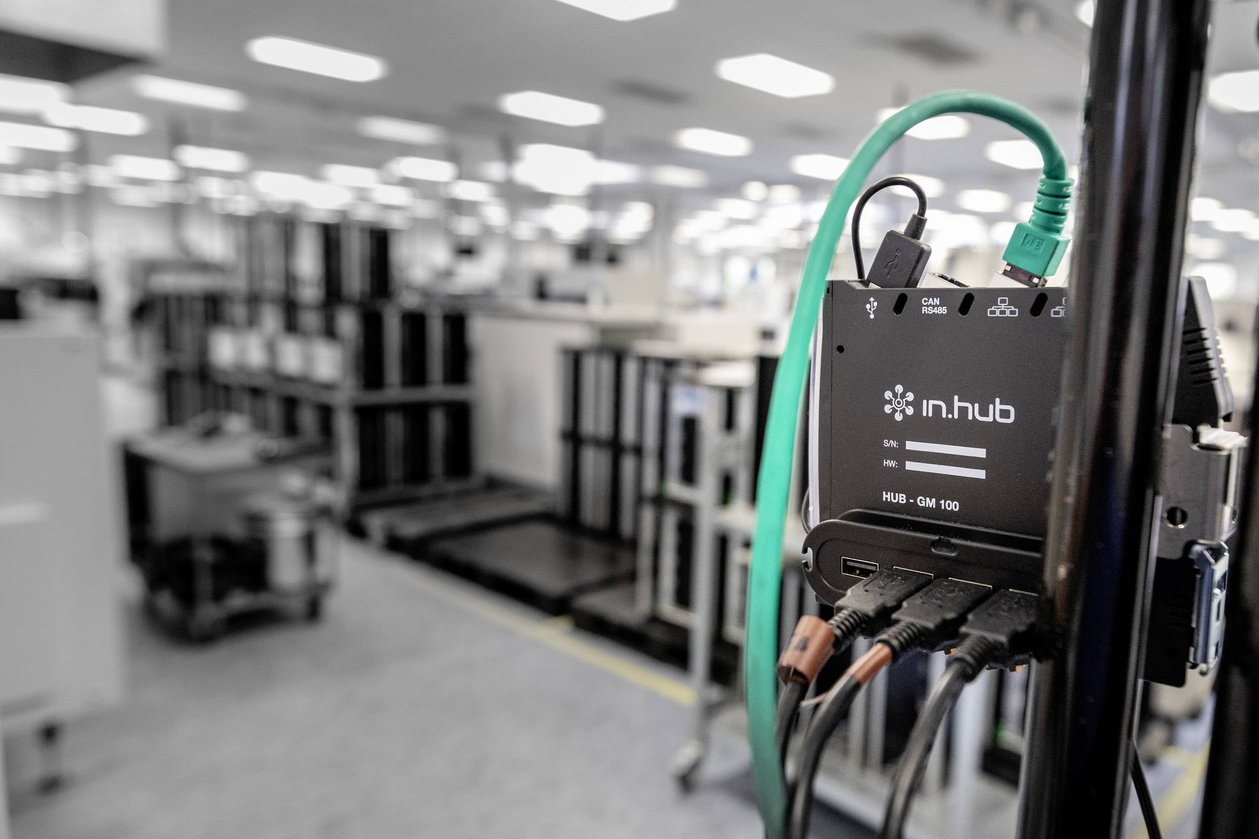 Un hub noir avec des câbles verts et noirs relie des appareils dans un environnement industriel. Des machines floues sont visibles en arrière-plan.