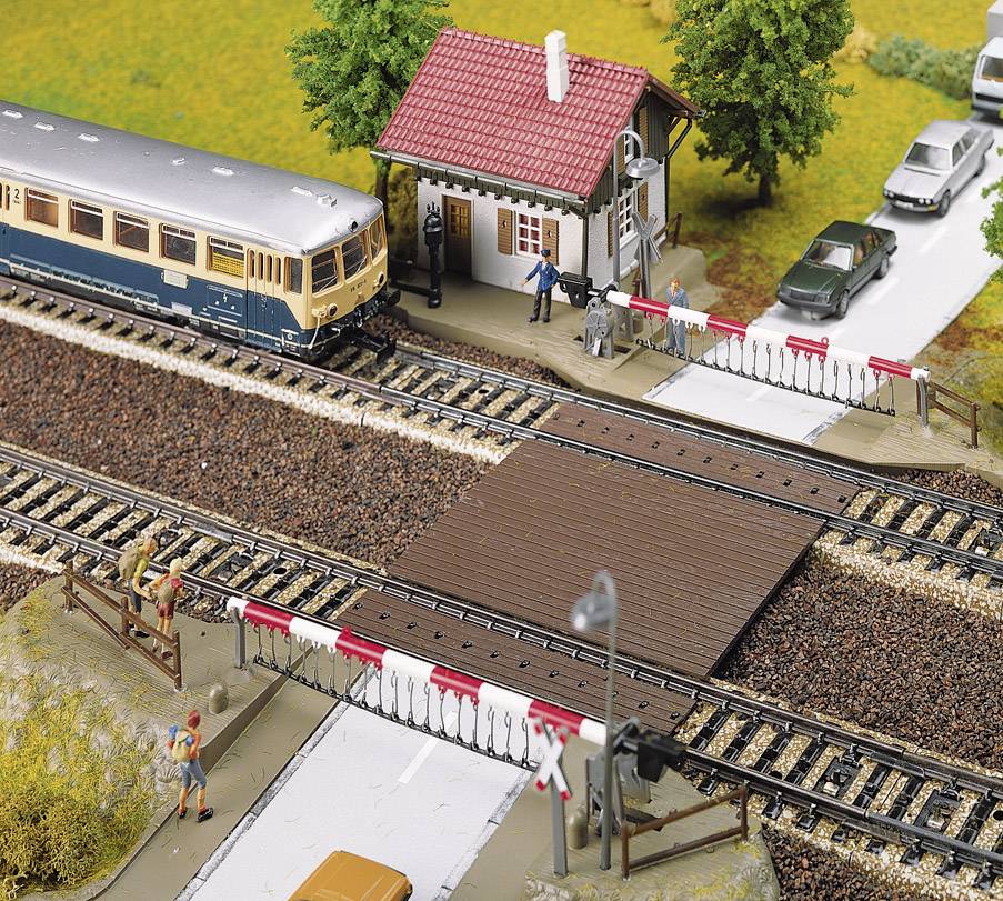 Un train miniature passe à un passage à niveau où des personnes sont postées et regardent. Des voitures attendent pendant que le train traverse.