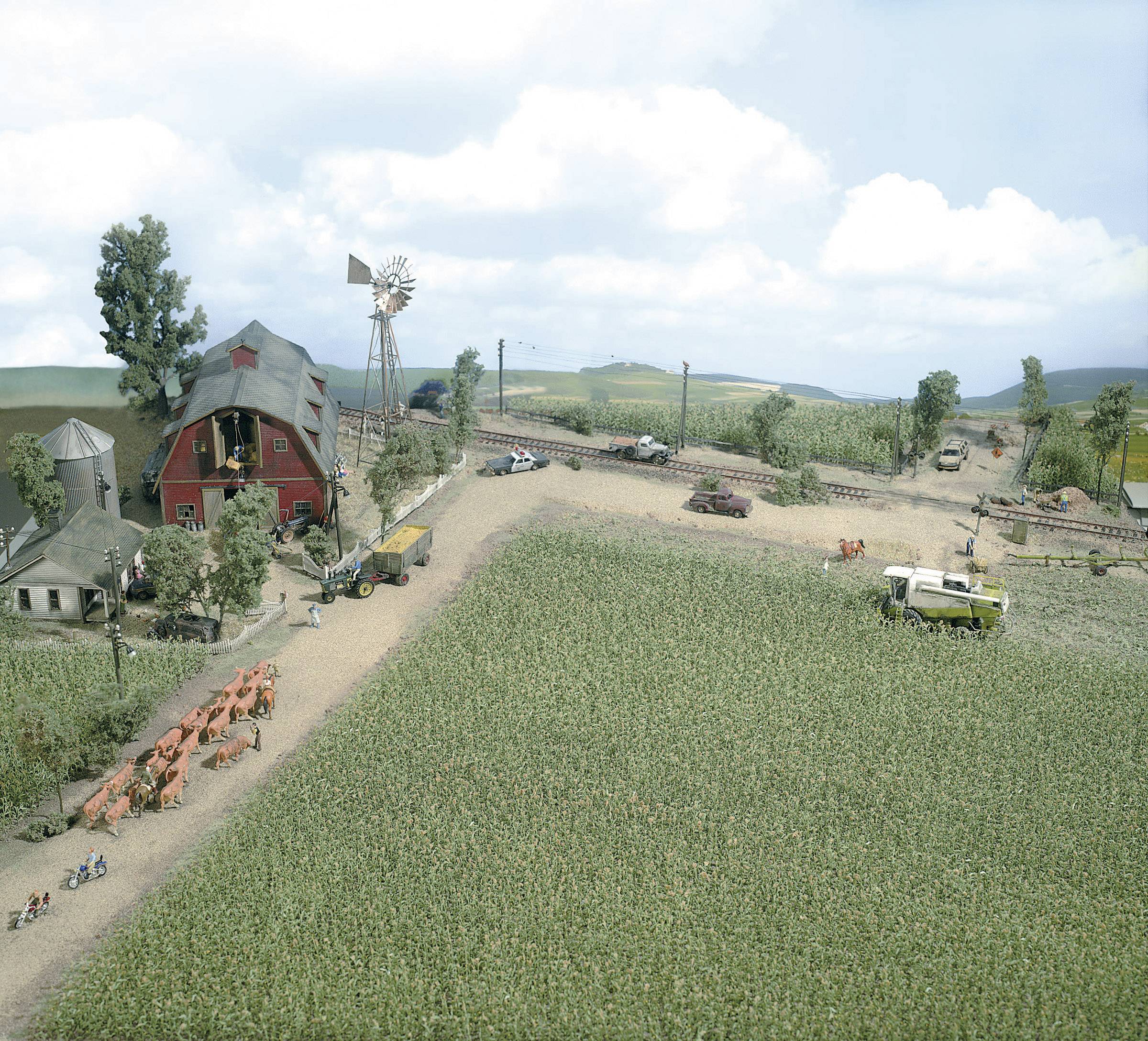 Vue aérienne d'une ferme avec une grange rouge, un moulin à vent et des champs. Des vaches paissent, et des machines agricoles sont visibles.
