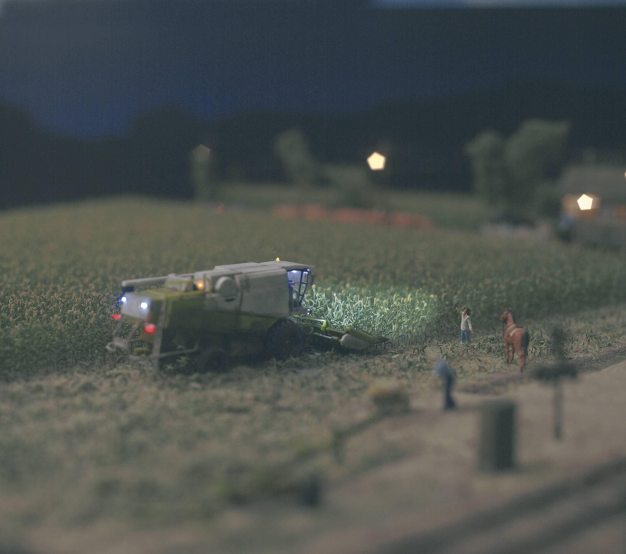 Une moissonneuse-batteuse travaille la nuit dans un champ. Deux personnes se tiennent au bord du champ, à côté d'elles un cheval. Des lumières sont visibles en arrière-plan.