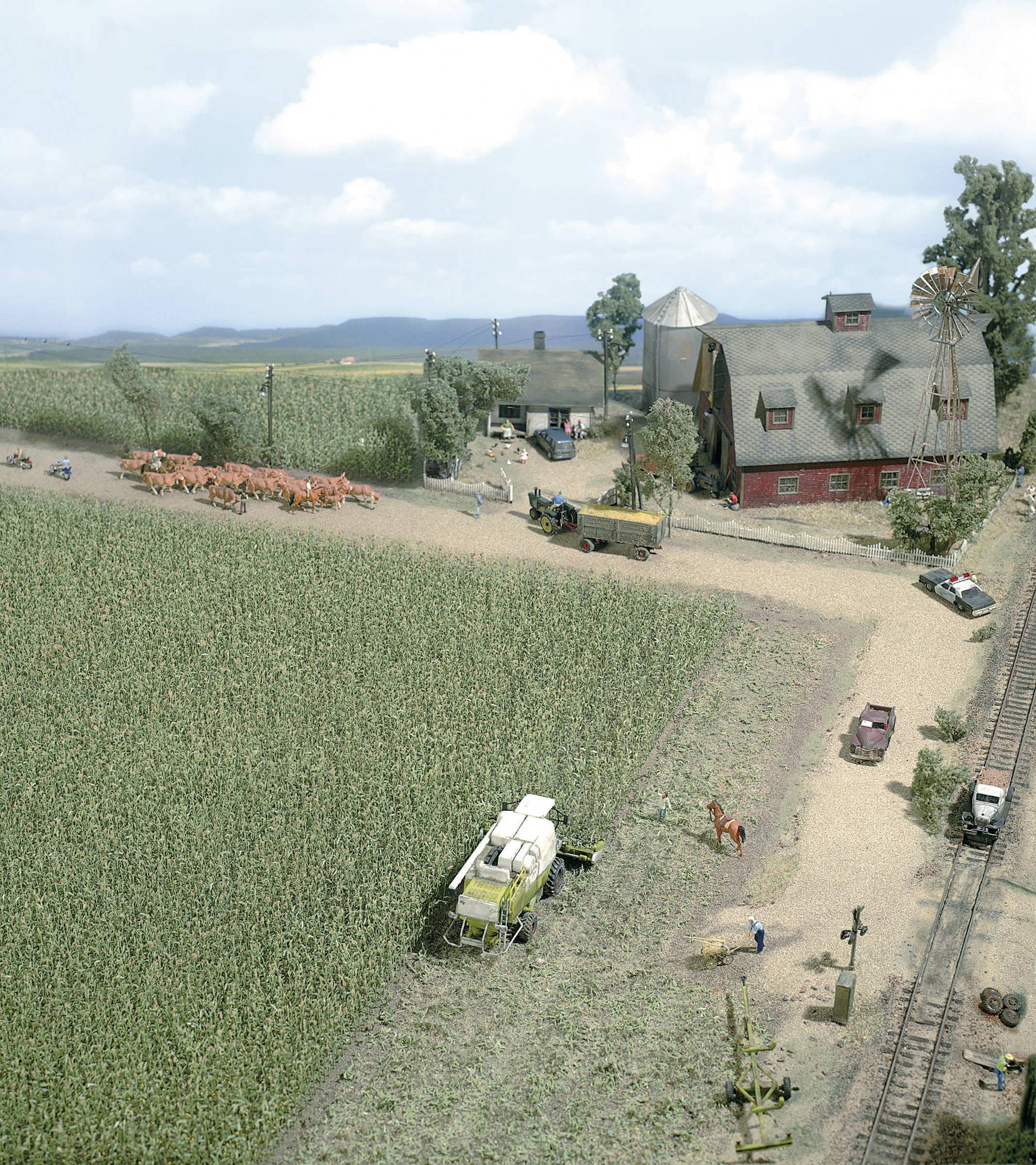 Paysage miniature d'une ferme avec un bâtiment rouge, des vaches sur un chemin de campagne, une moissonneuse-batteuse et des véhicules agricoles.